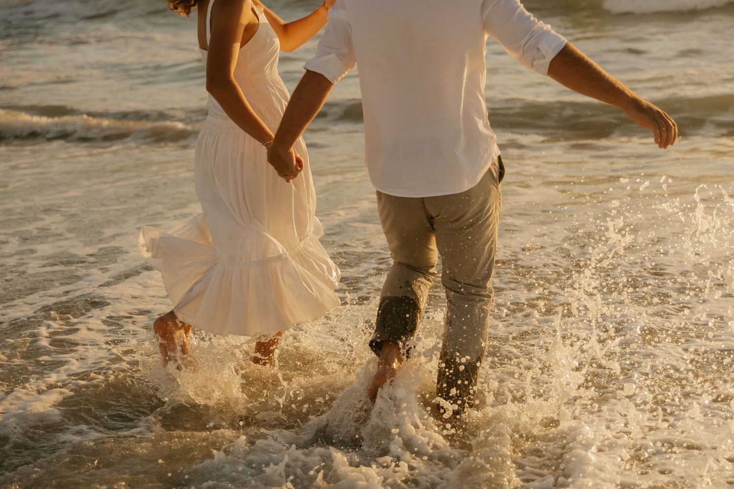 Couple splashing through ocean waves at sunset wearing white and beige coastal attire.