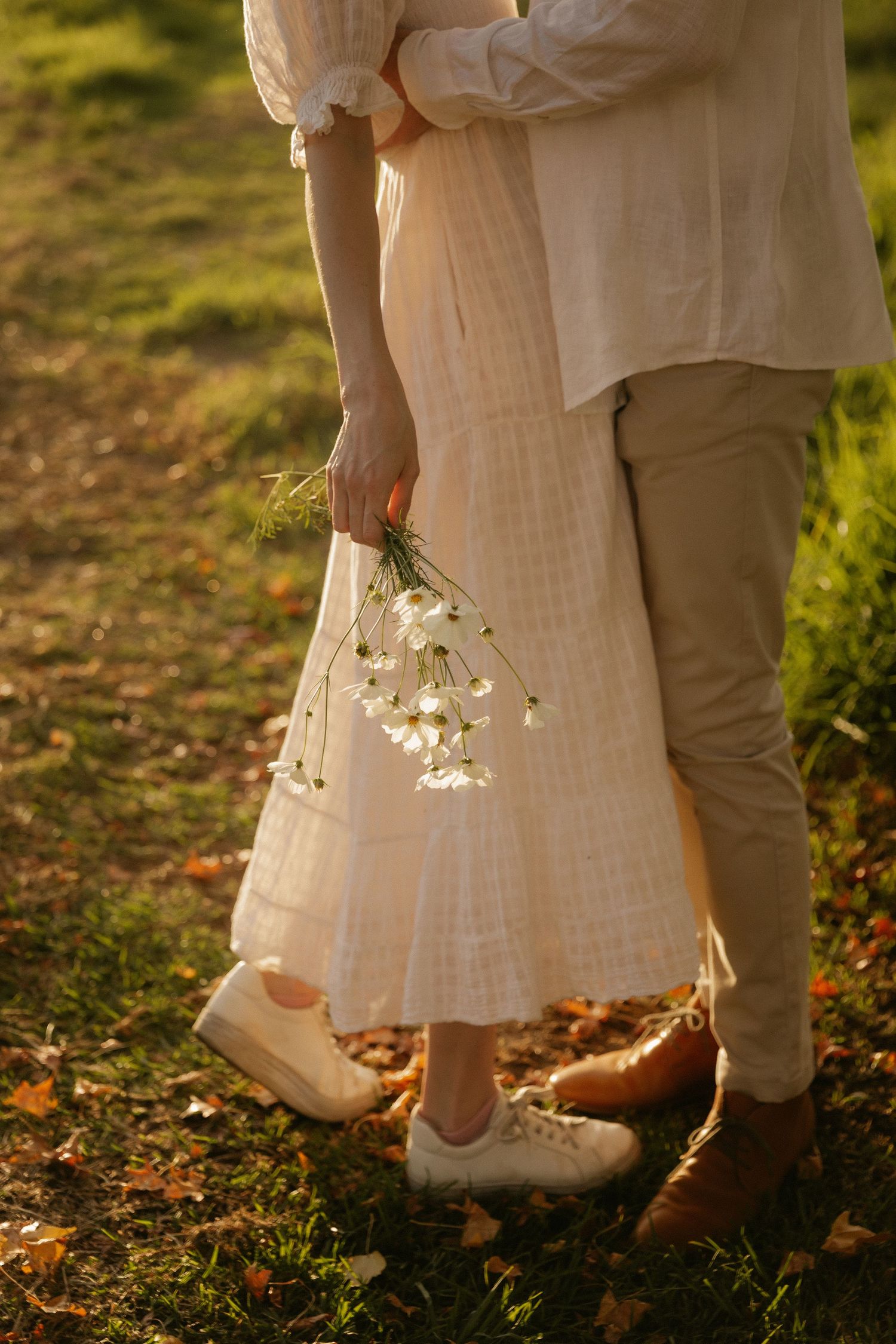 Two people in light colored clothing stand close together holding white flowers in golden sunlight on grass with autumn leaves.