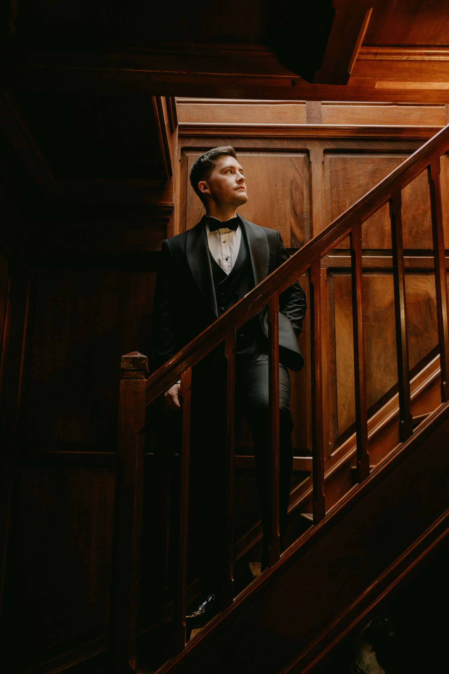 A person in formal attire stands on wooden stairs in a dark, moody setting with dramatic lighting.
