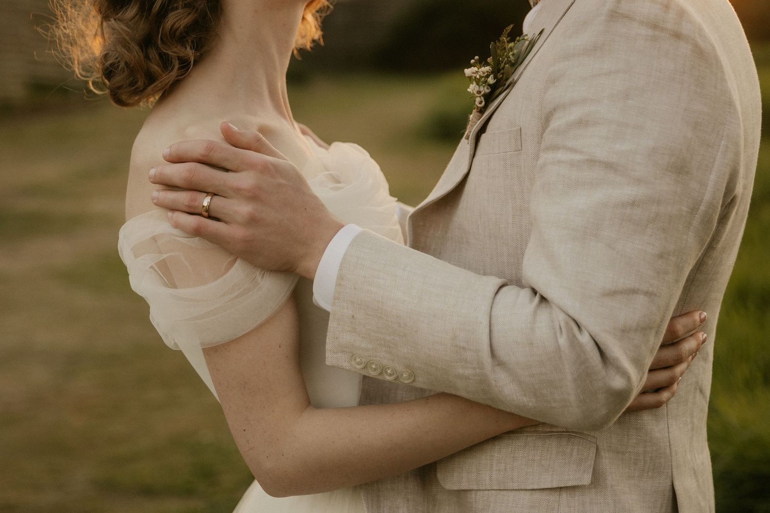 A close-up shot of a couple embracing in soft sunlight, wearing light-colored wedding attire and a sparkling ring.