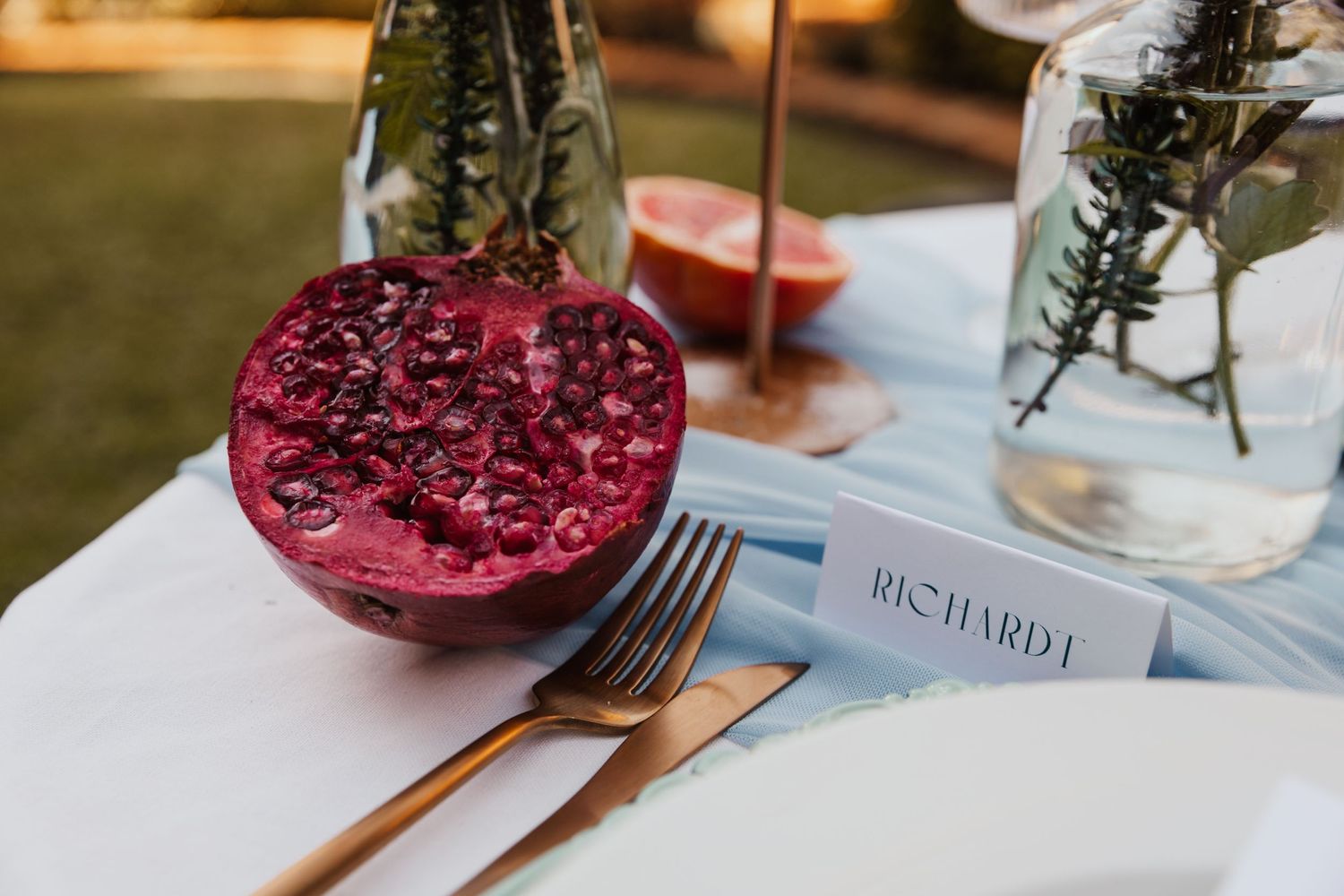 Place setting with red pomegranate, cutlery, and name card on white tablecloth.
