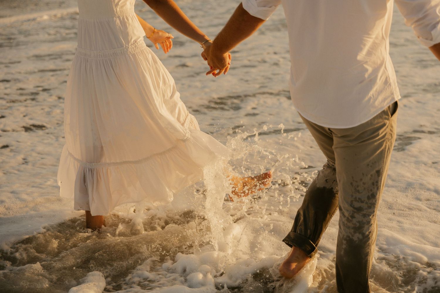 A couple in white outfits holds hands while walking on a sandy beach during sunset.