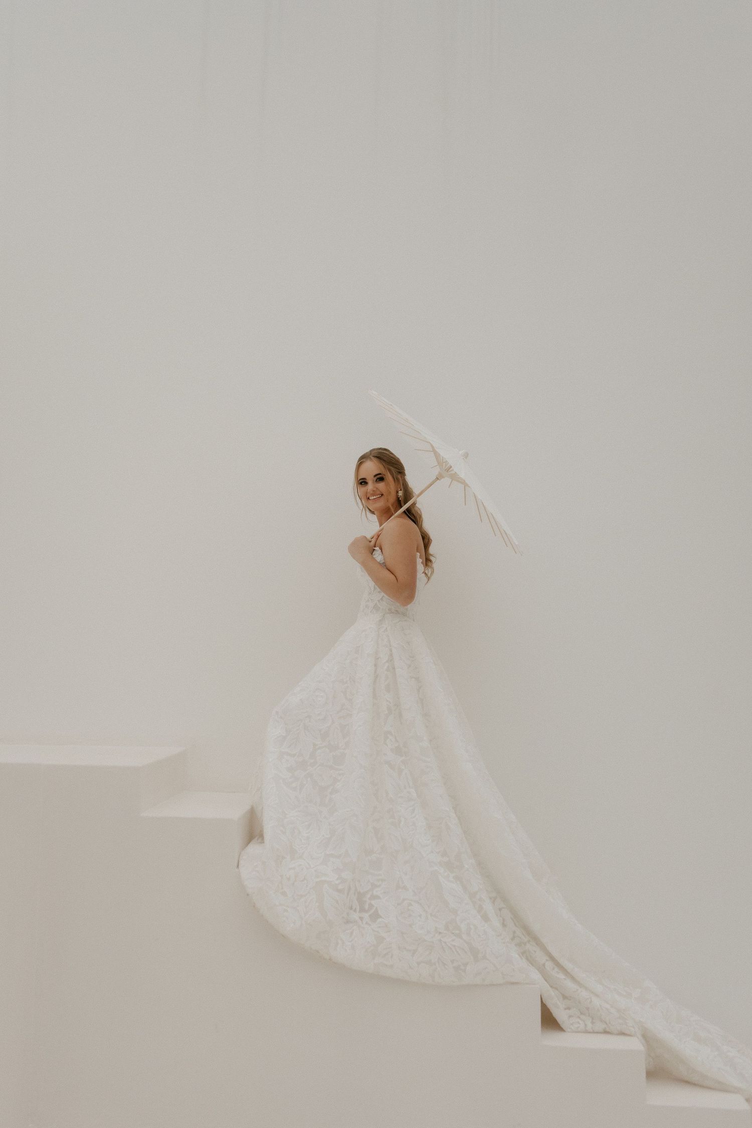 Bride in lace ball gown with long train holds white parasol on white steps, smiling over shoulder.