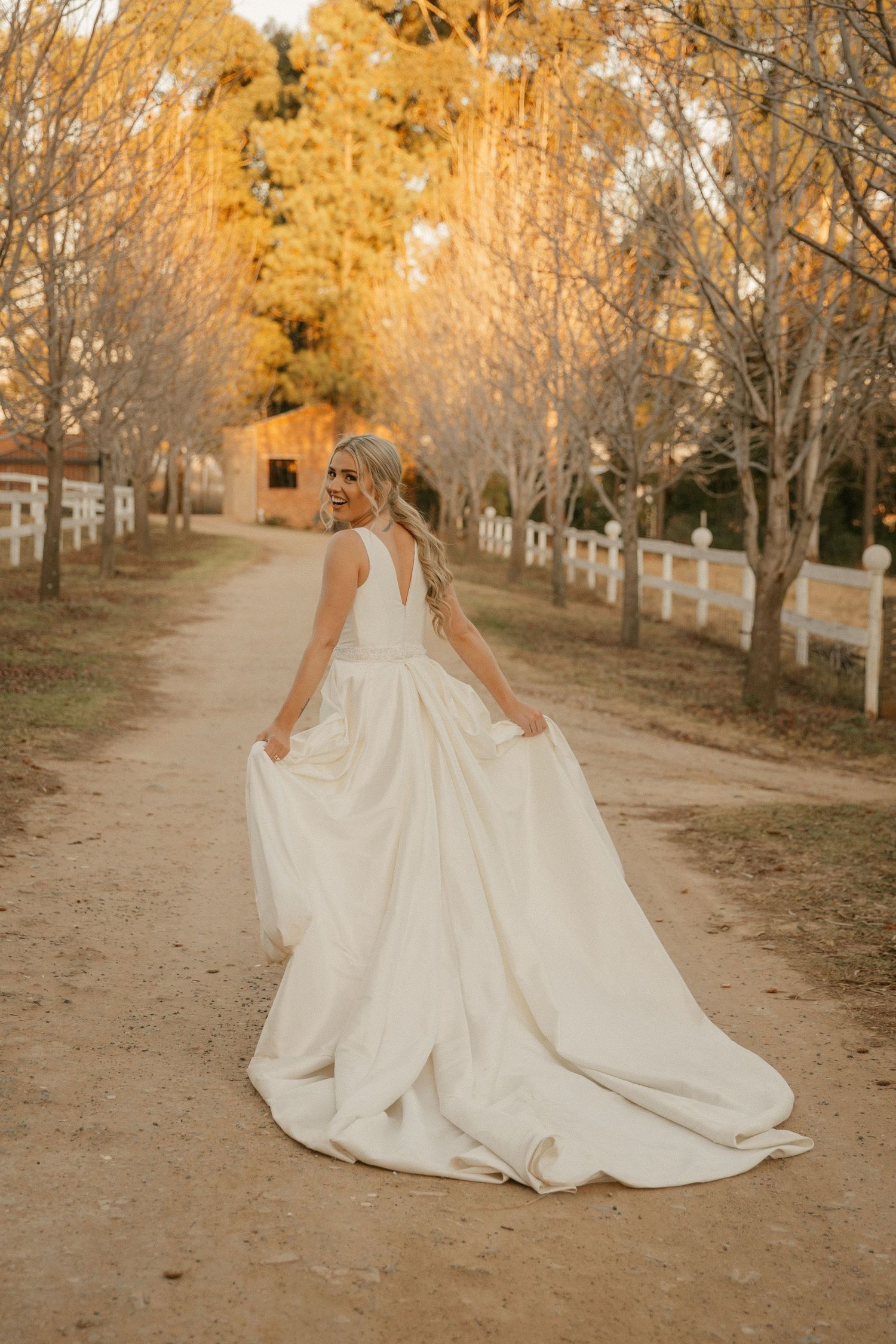 Beautiful fall wedding dress photography showing flowing white gown against a rustic dirt path lined with autumn trees.