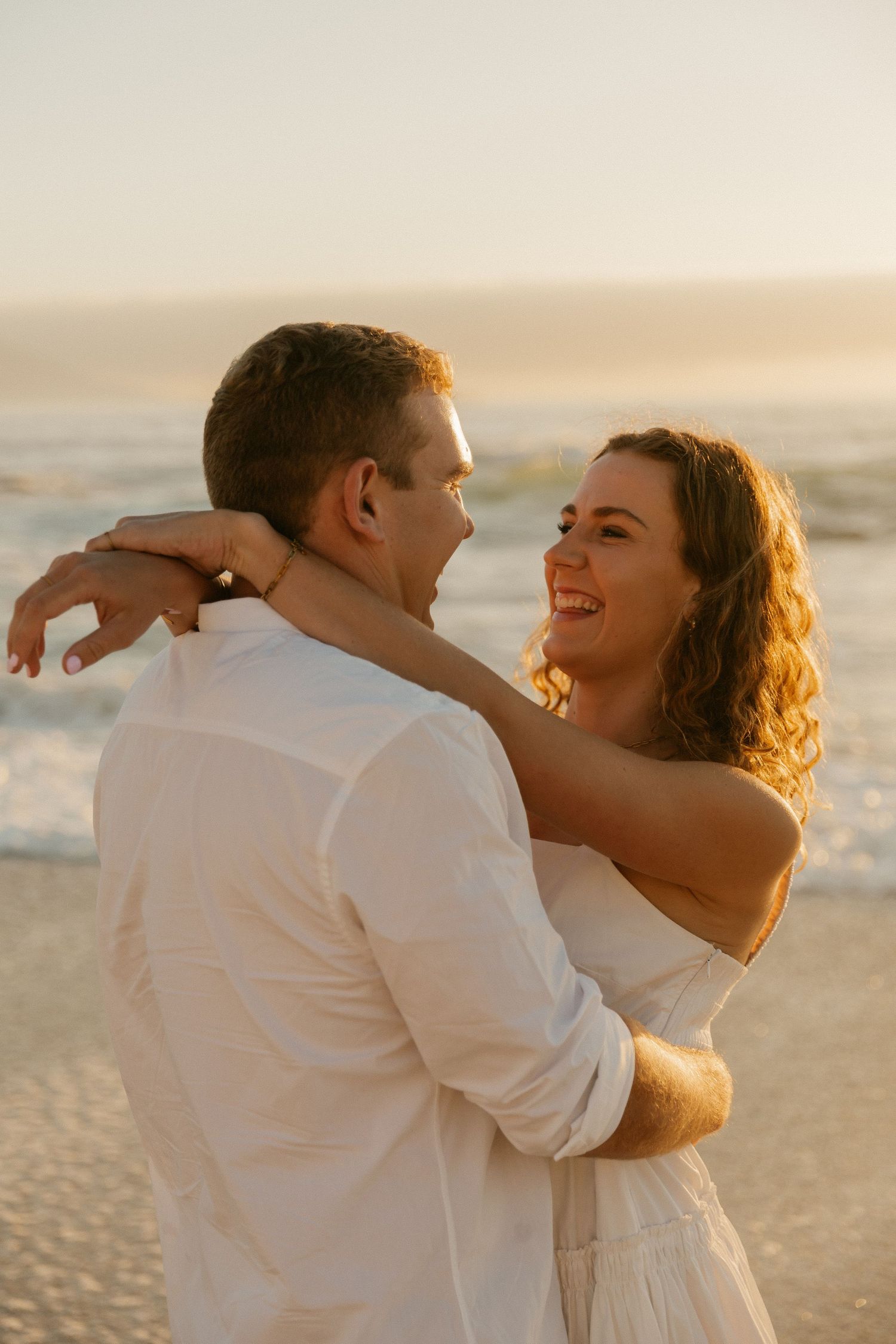 A romantic couple embraces on the beach during sunset wearing matching white outfits.