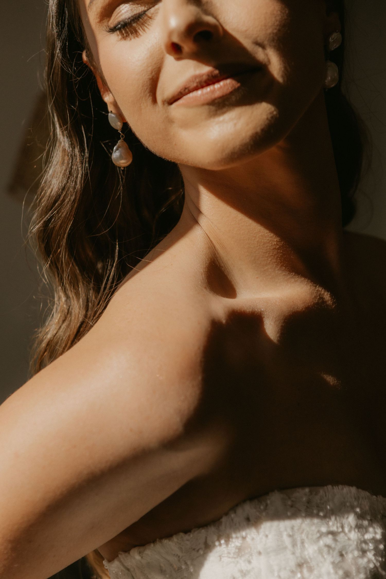 A bride in a strapless lace gown enjoys warm sunlight, wearing elegant drop earrings with eyes closed.