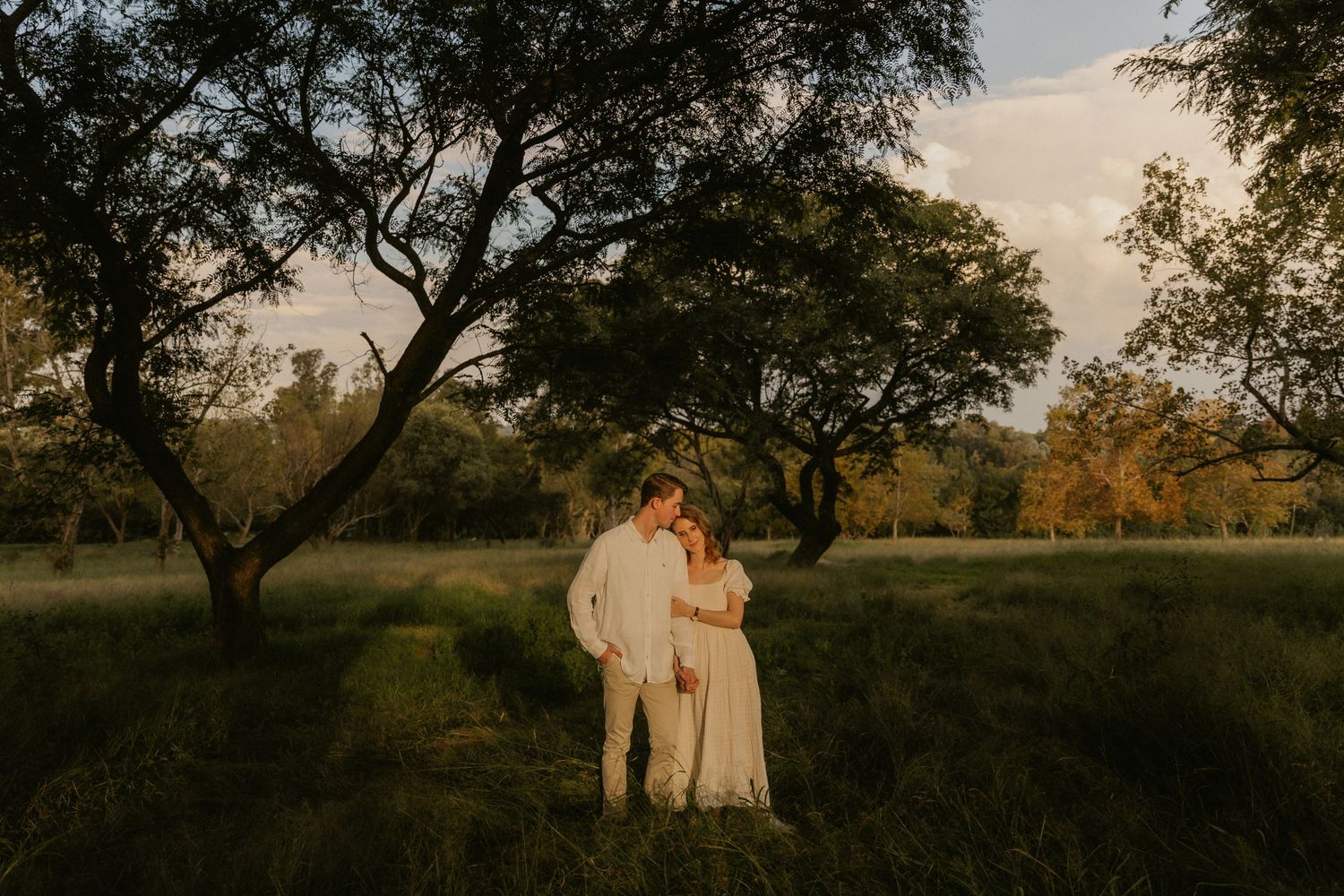 A couple in white outfits embraces during sunset in a romantic outdoor photoshoot at a grassy field.