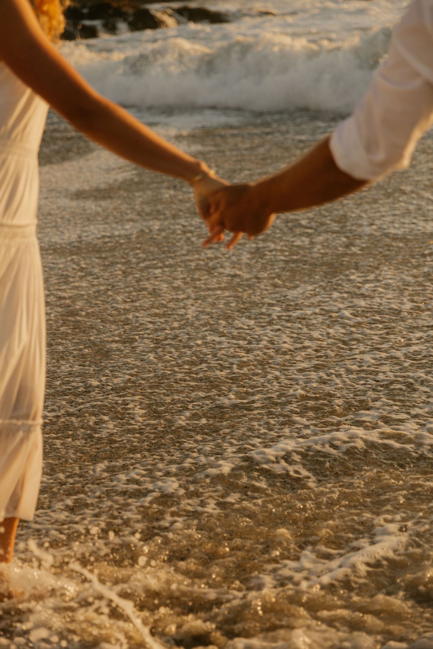 Close-up of intertwined hands as ocean waves wash over the sandy shore at sunset.