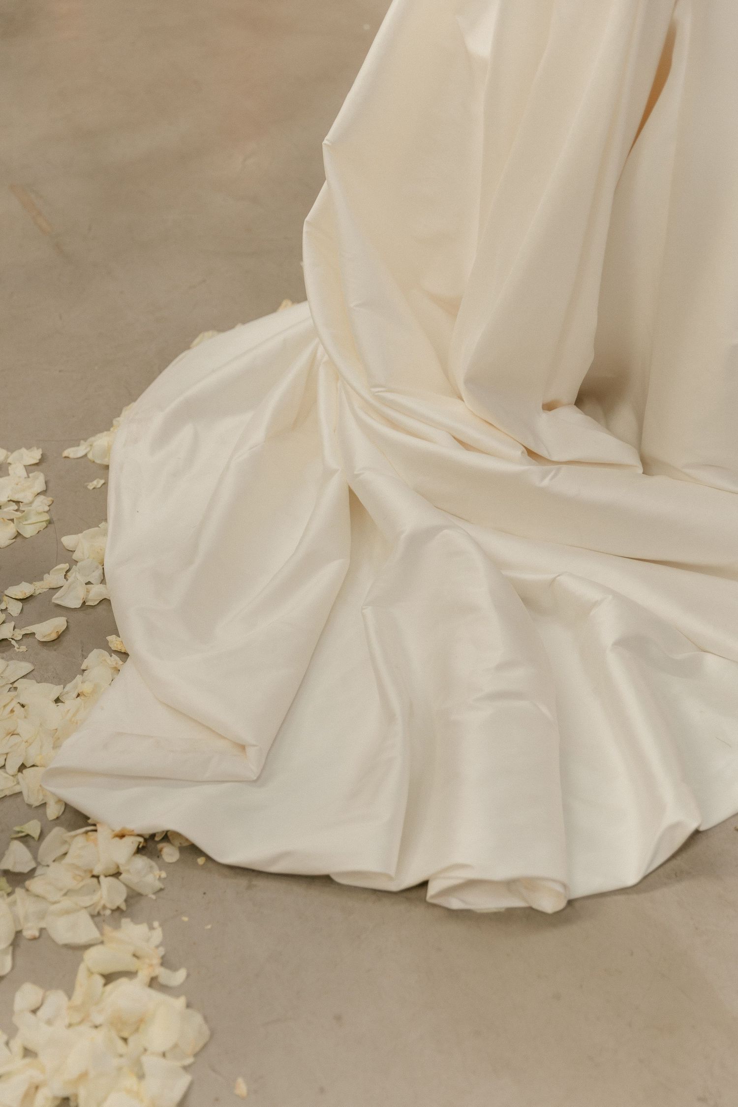 Flowing ivory wedding dress hem with delicate flower petals scattered on concrete floor.