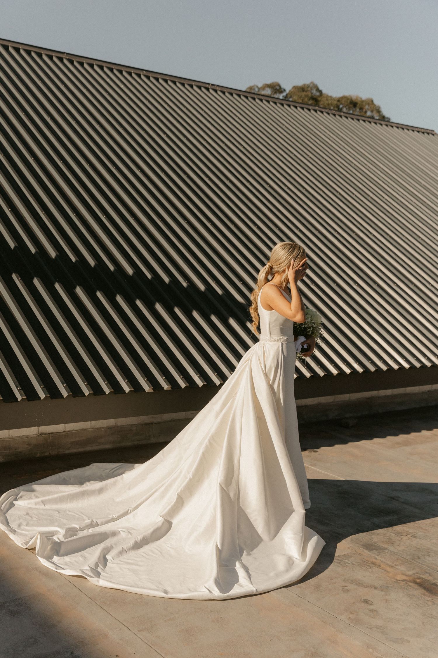 A wedding dress train flows elegantly across a concrete surface against a curved metal roof backdrop.
