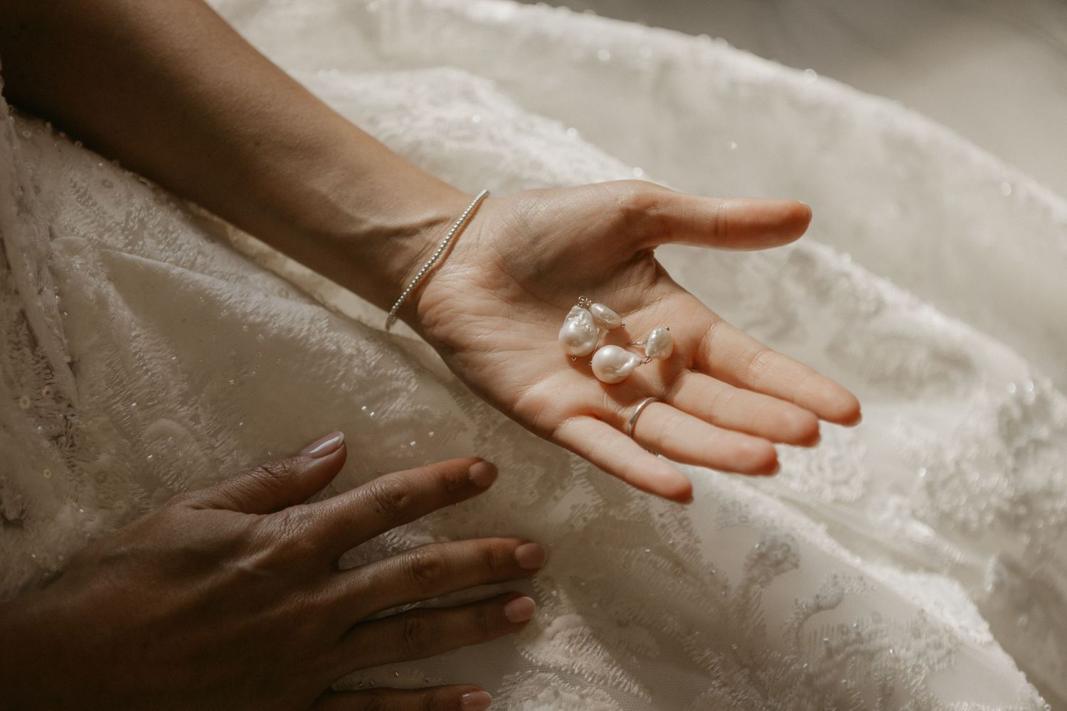 Bride holding pearl earrings in her open palm against a white lace wedding dress.