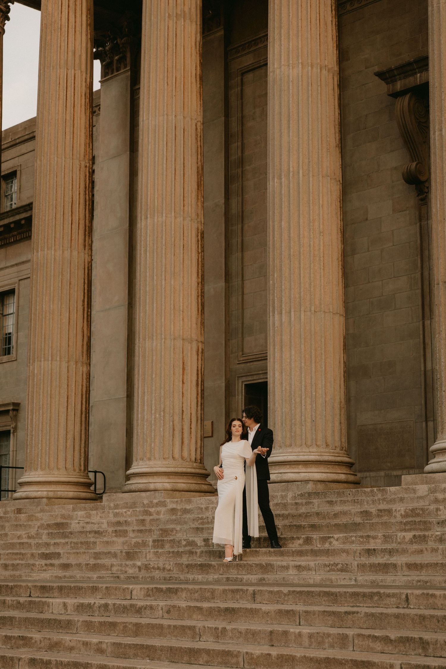 A couple stands between grand stone columns on the steps of a neoclassical building with warm lighting.