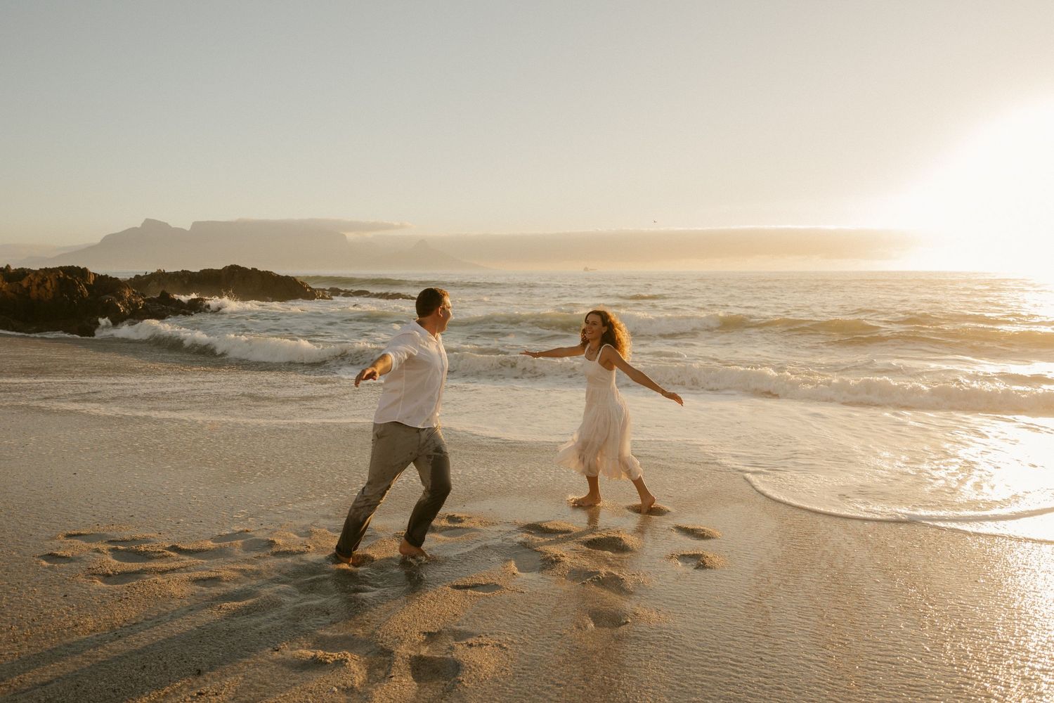 A couple playfully dances on a sandy beach at sunset with crashing waves in the background.