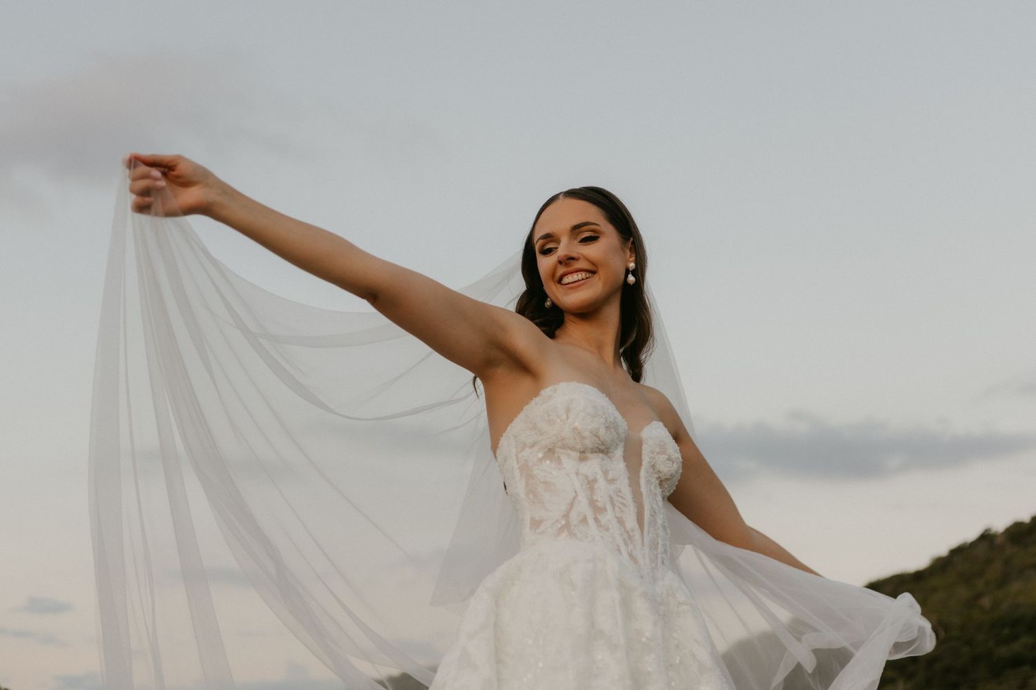 Radiant bride in lace gown twirling her veil against a soft evening sky backdrop.