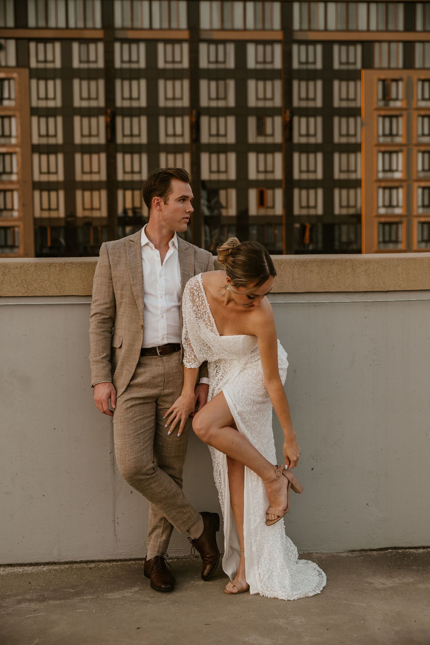 A couple in wedding attire sits together on a rooftop ledge with brick buildings in the background.