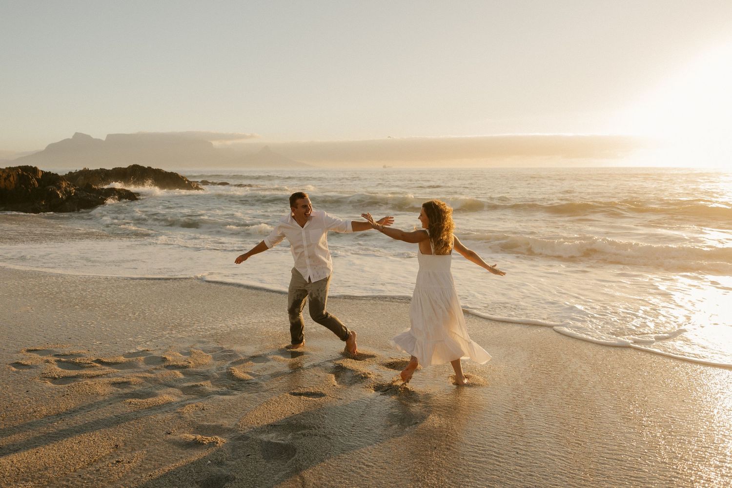 A couple dances playfully together on a beach at golden hour with waves crashing behind them.