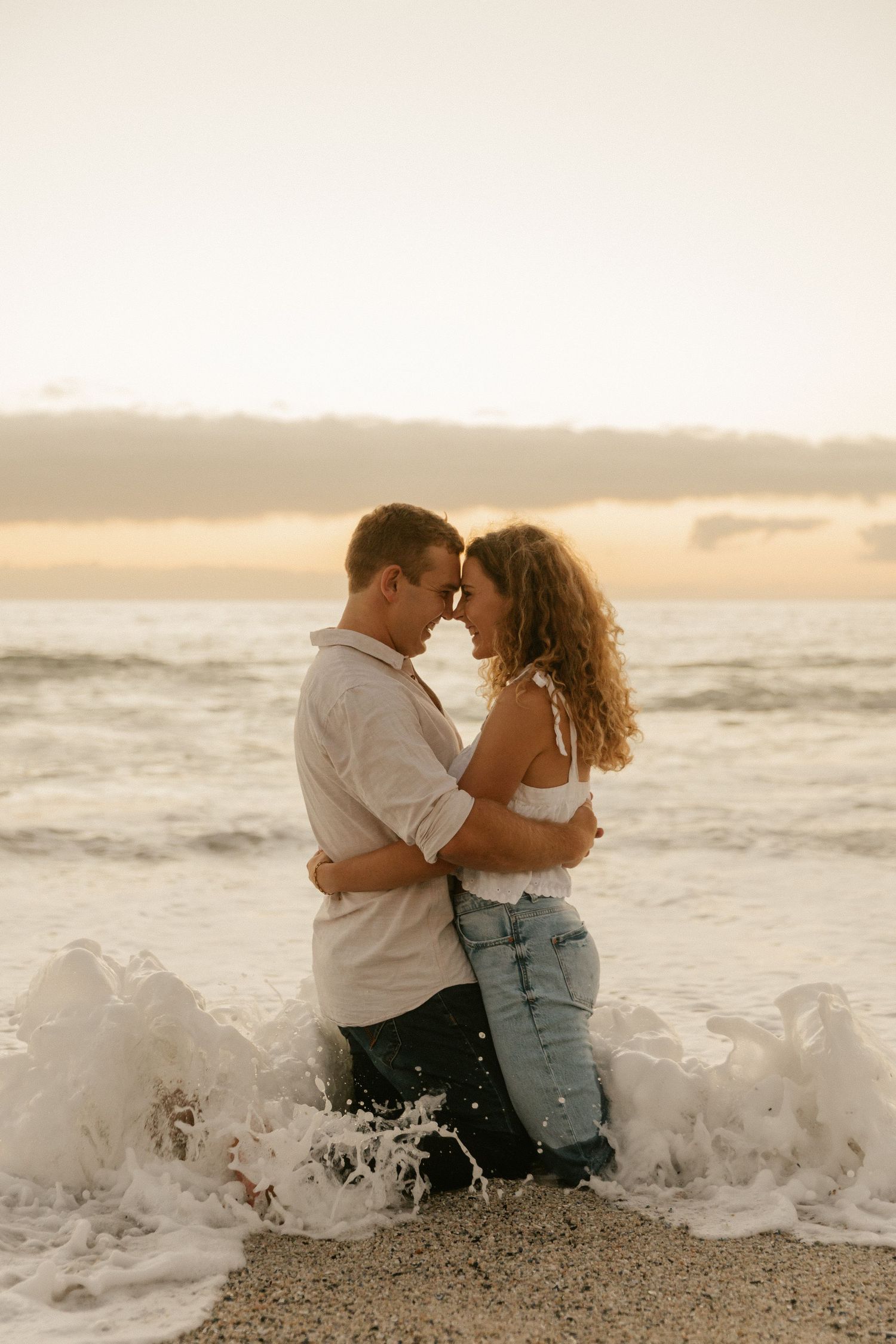 Couple embraces in the surf as ocean waves crash around them during sunset.