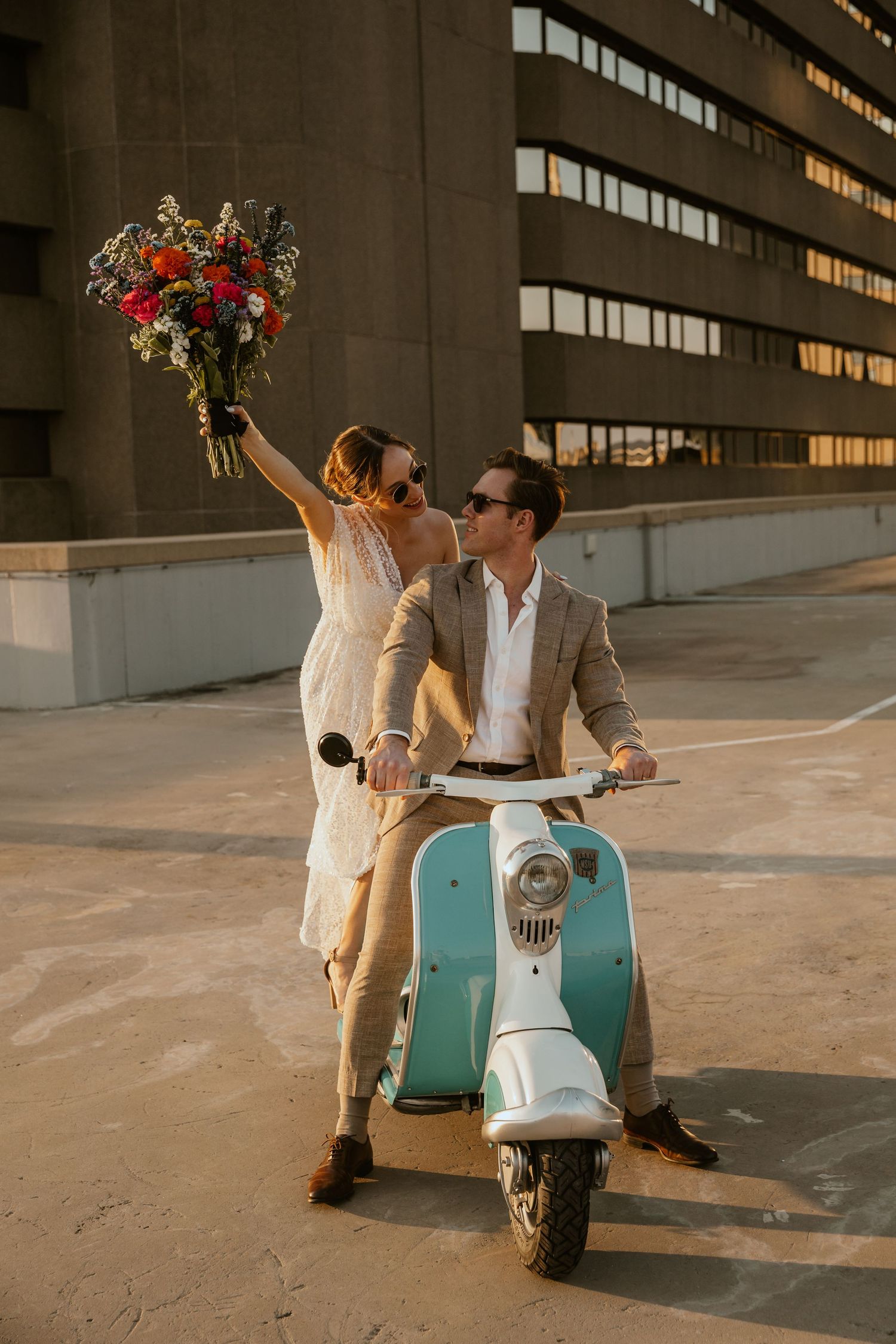 A couple on a mint scooter holds up a vibrant red and orange wedding bouquet against an urban backdrop.