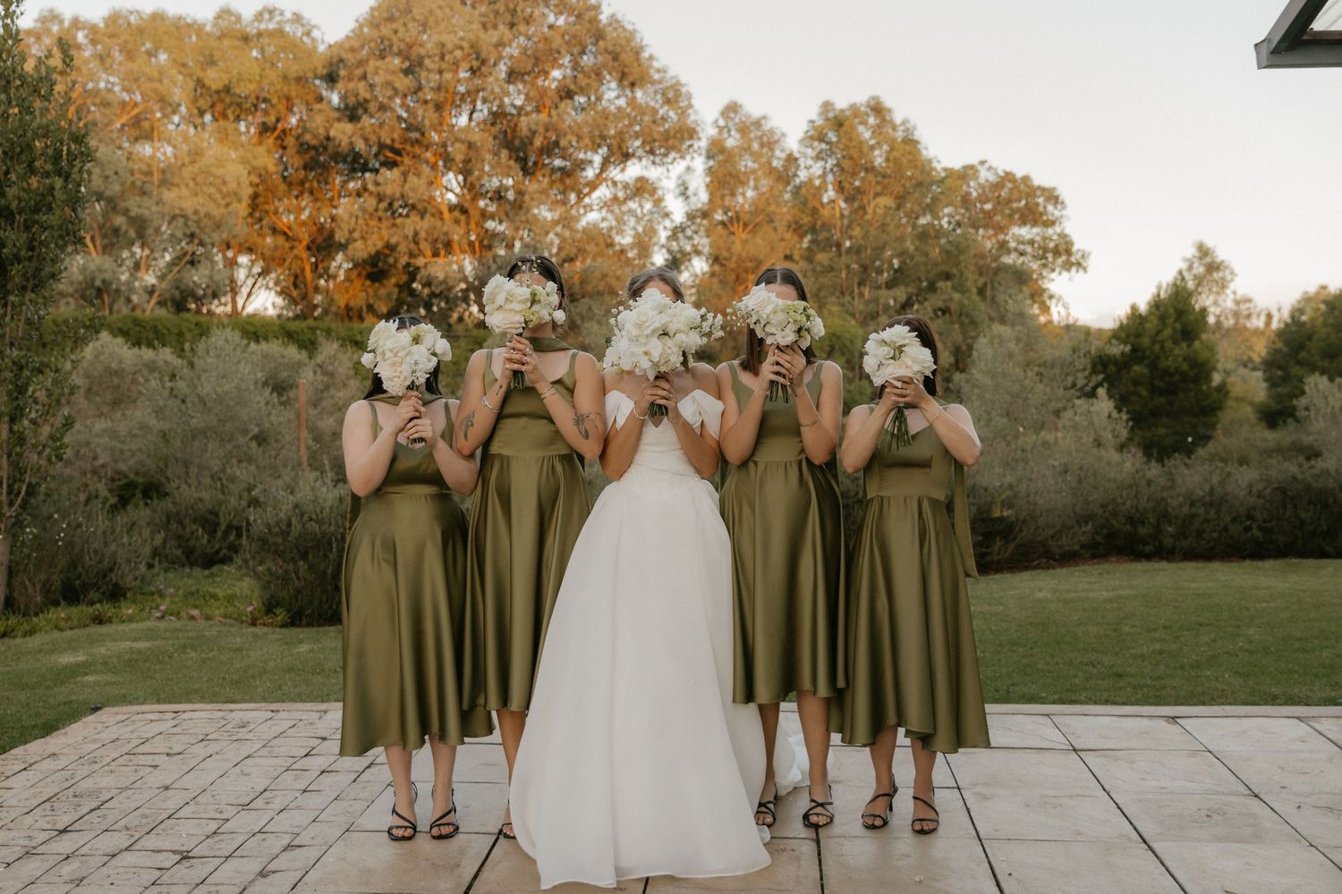 Wedding party in olive green dresses and white gown pose together in an autumn outdoor setting at sunset.