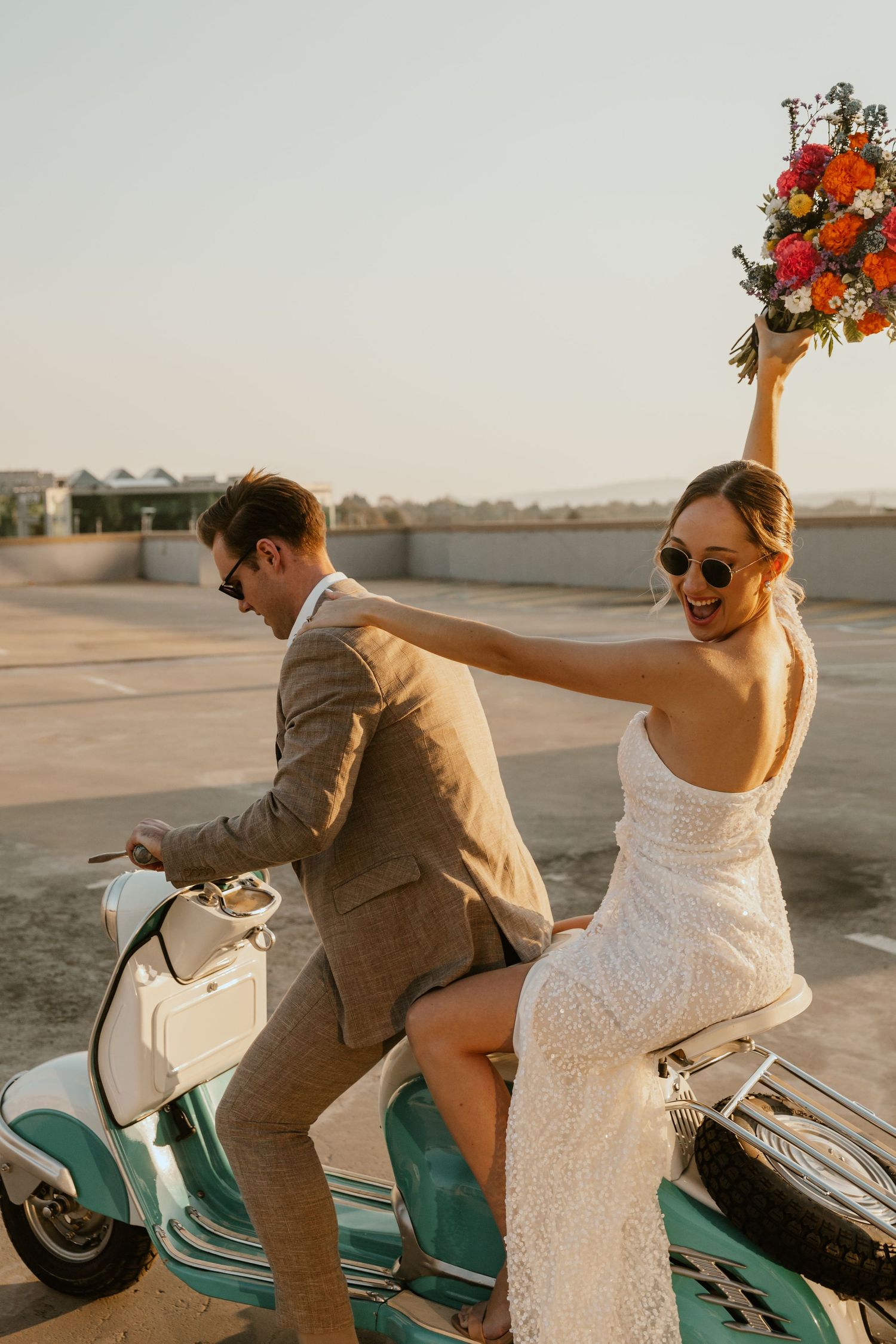 Newlyweds ride a vintage mint-colored Vespa scooter on a rooftop parking area during sunset.