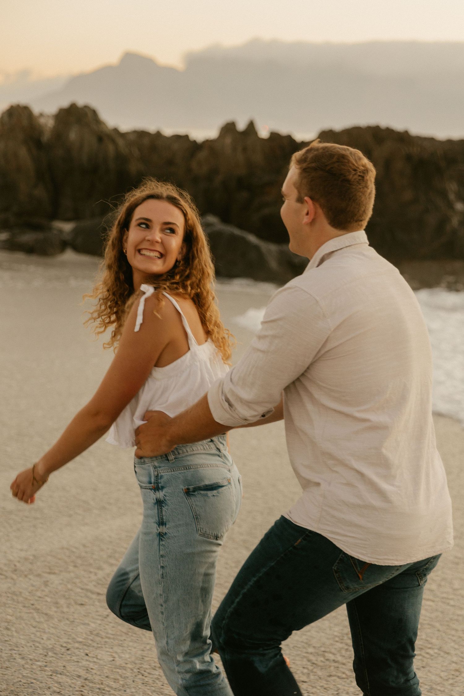 Couple dances together on the beach with rocky cliffs and sunset in the background.