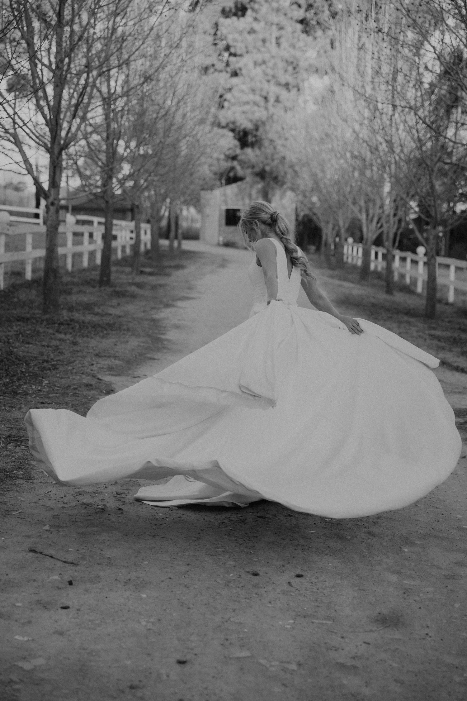 A figure in a flowing white wedding dress twirls gracefully on a tree-lined path in black and white photography.