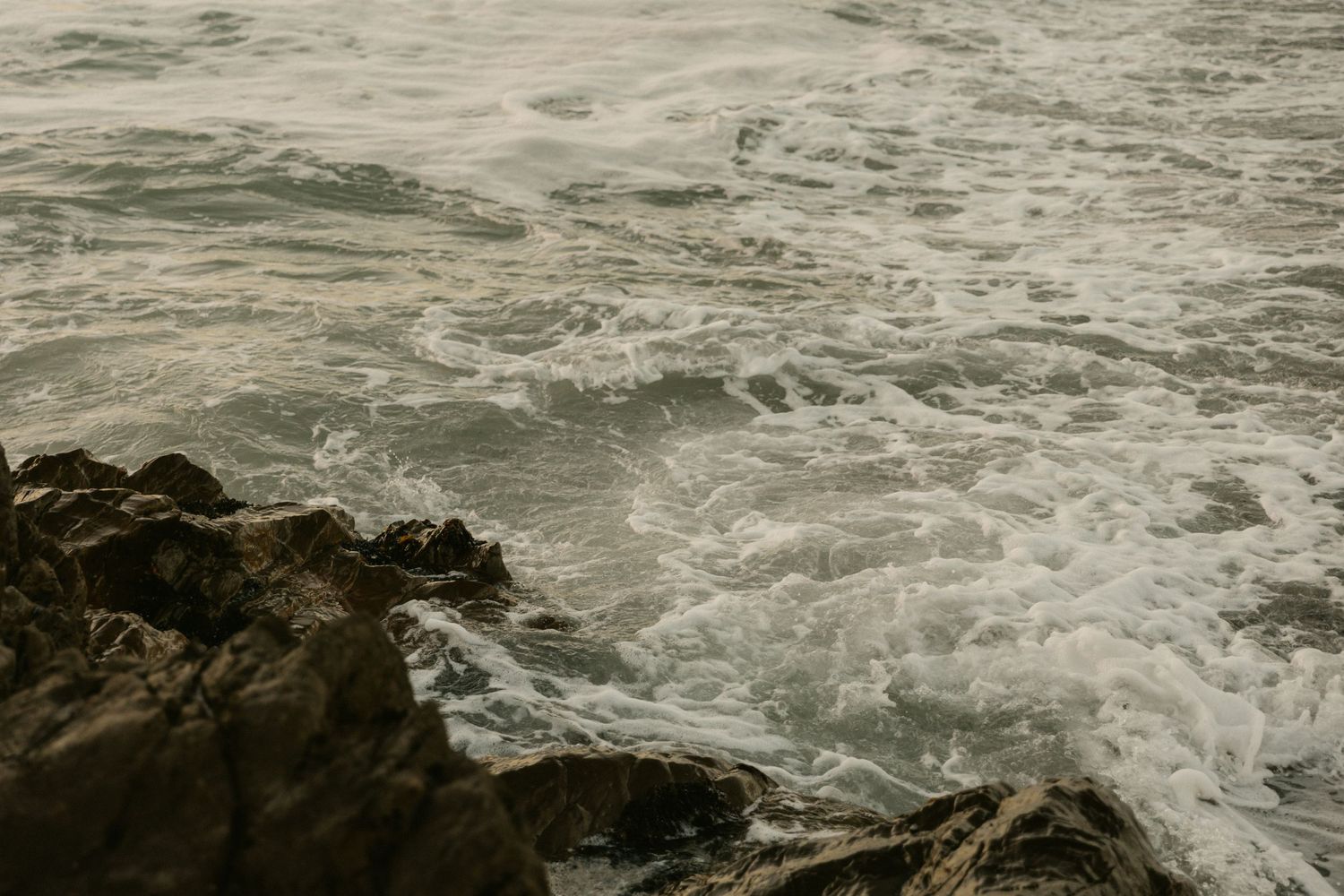 Ocean waves crash against dark rocky coastline during sunset.