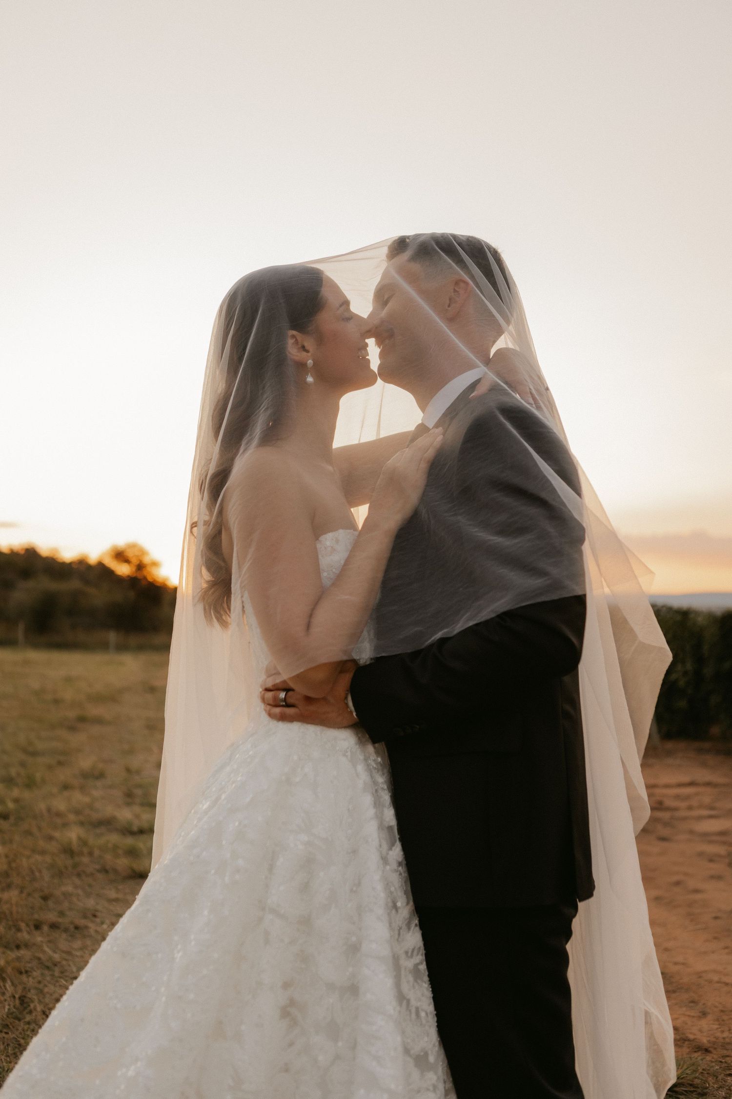 Bride and groom share a romantic moment under a flowing veil at sunset in an outdoor field.