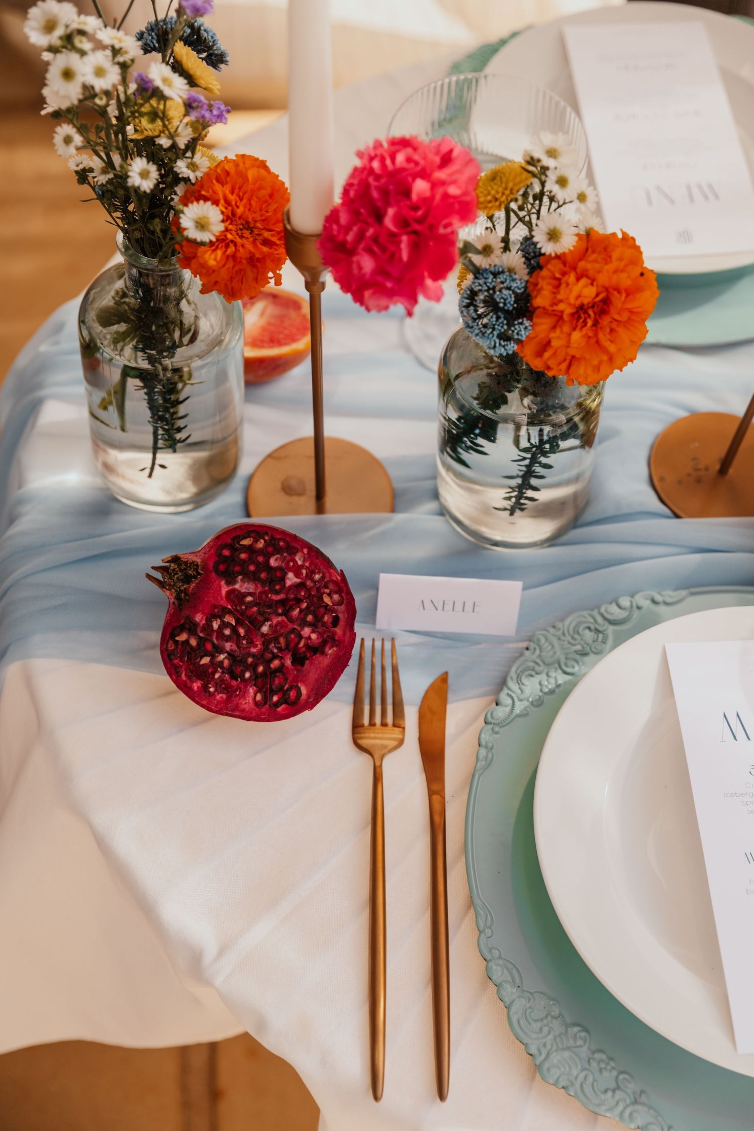 Wedding place setting featuring bright florals, pomegranate, and mint-colored plates with gold cutlery.