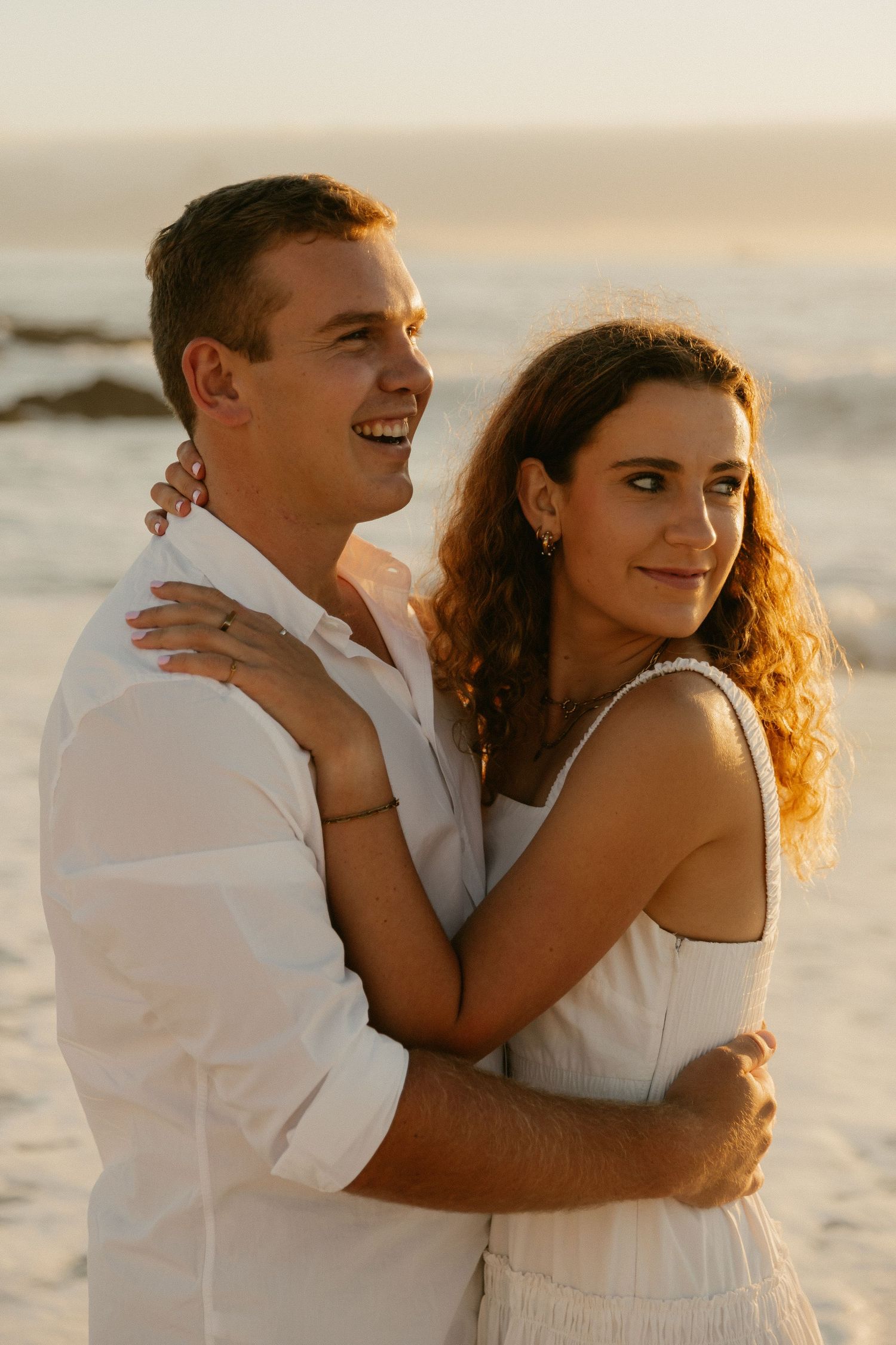 A couple in white clothing embraces while standing on a beach at sunset.