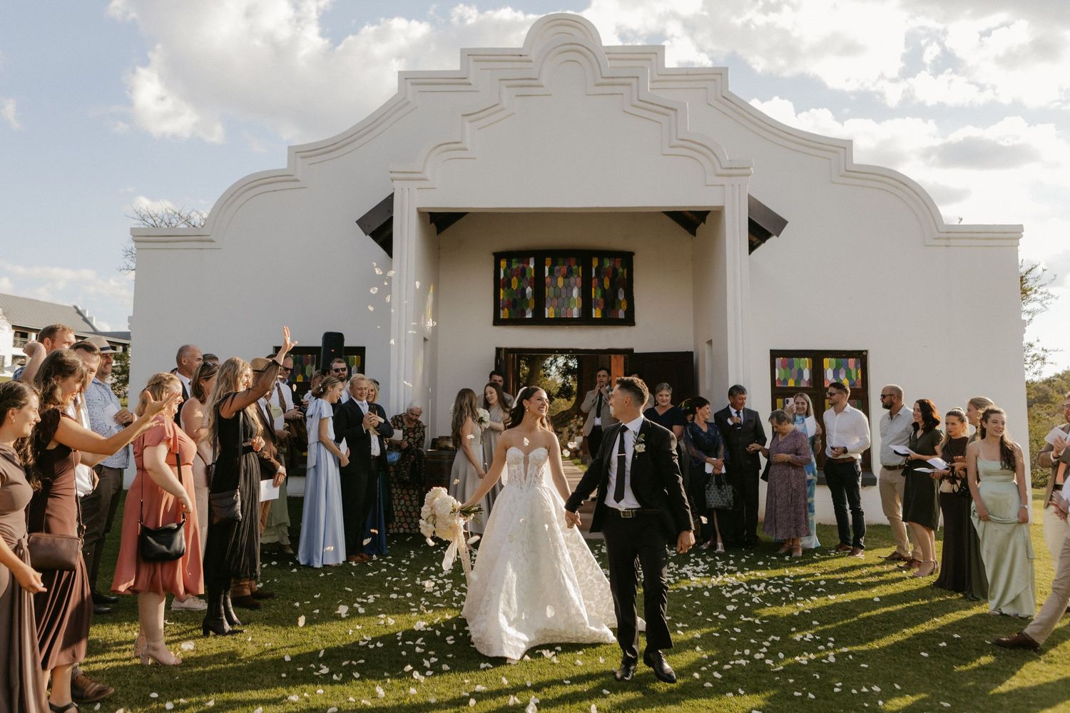 Bride and groom exit white chapel church as guests toss flower petals during outdoor wedding ceremony.