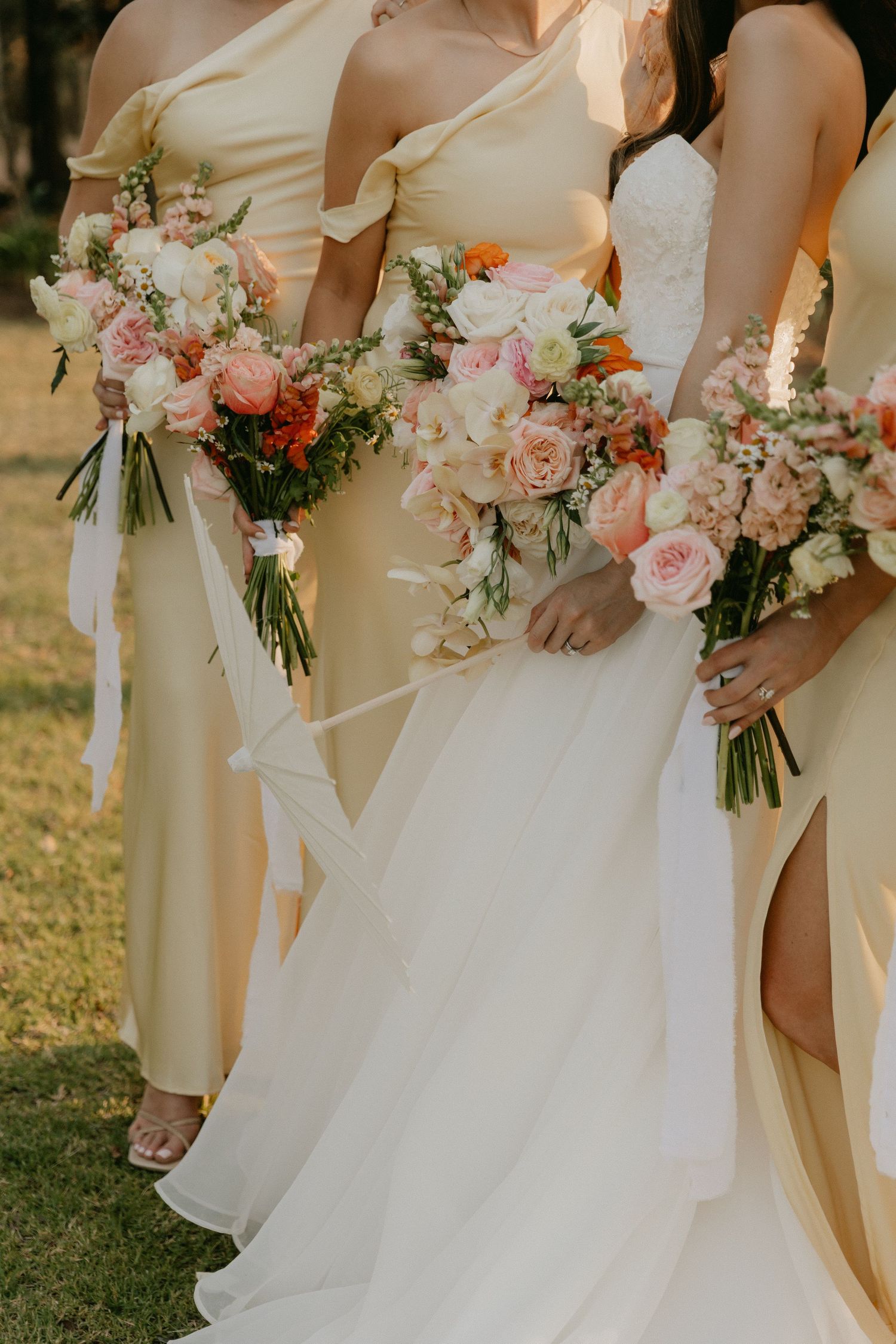 Bridesmaids in pale yellow dresses hold elegant pink and coral floral bouquets while standing on grass at an outdoor wedding.