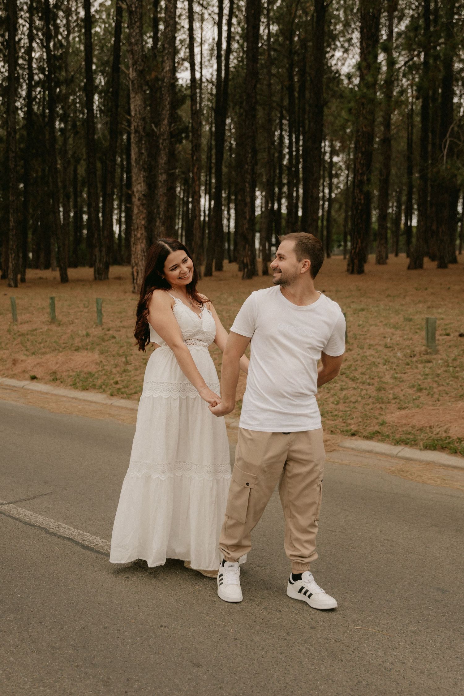 A couple in coordinated white outfits walk together and laugh on a tree-lined path in a romantic outdoor setting.