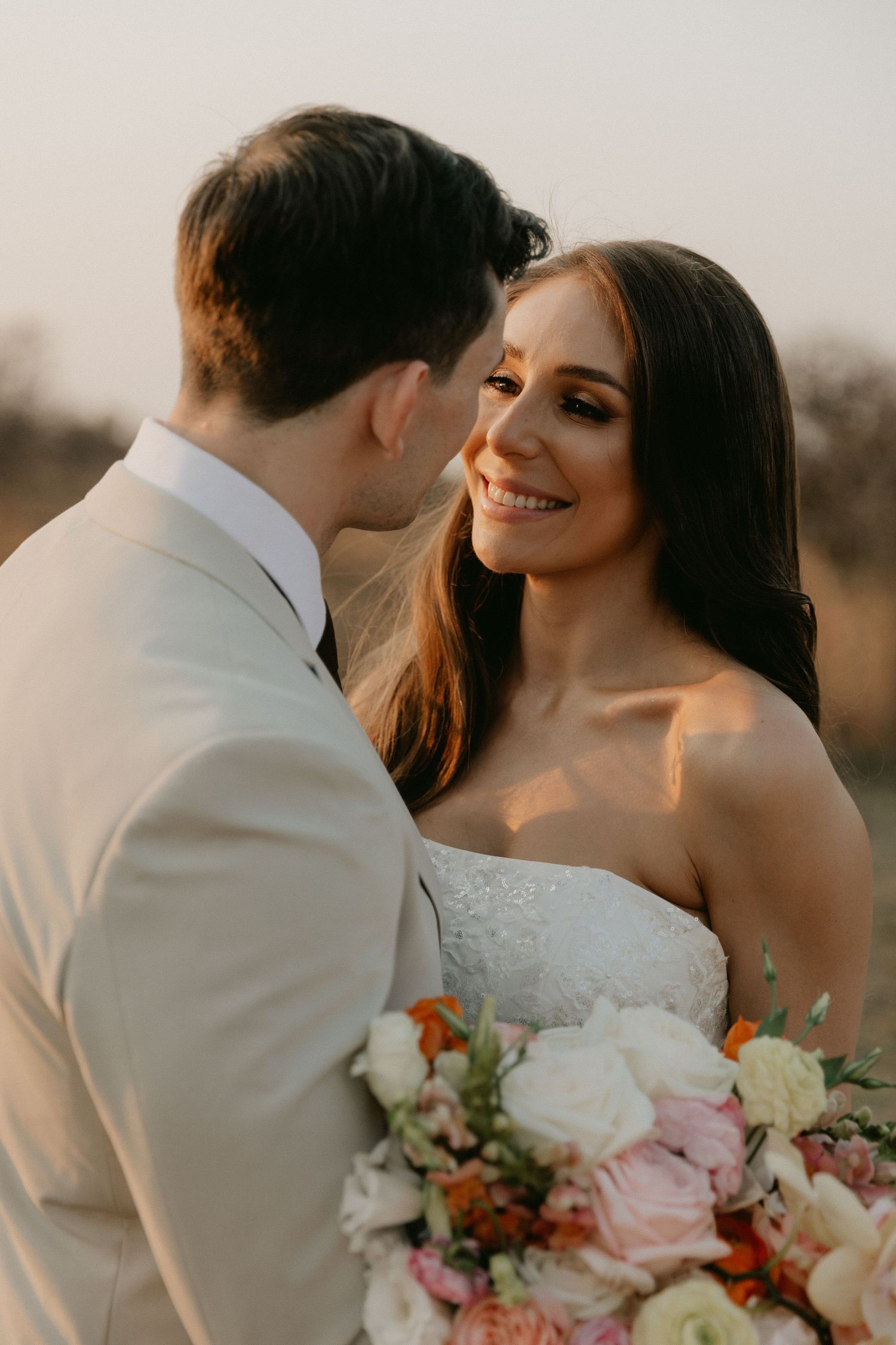 Newlywed couple shares romantic moments at sunset with pink and orange floral bouquet in foreground.