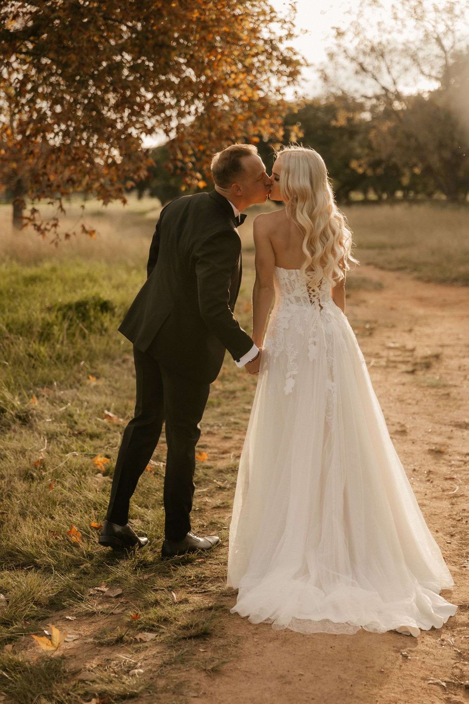 A couple in formal wedding attire share an intimate moment on a dirt path during golden hour under autumn trees.