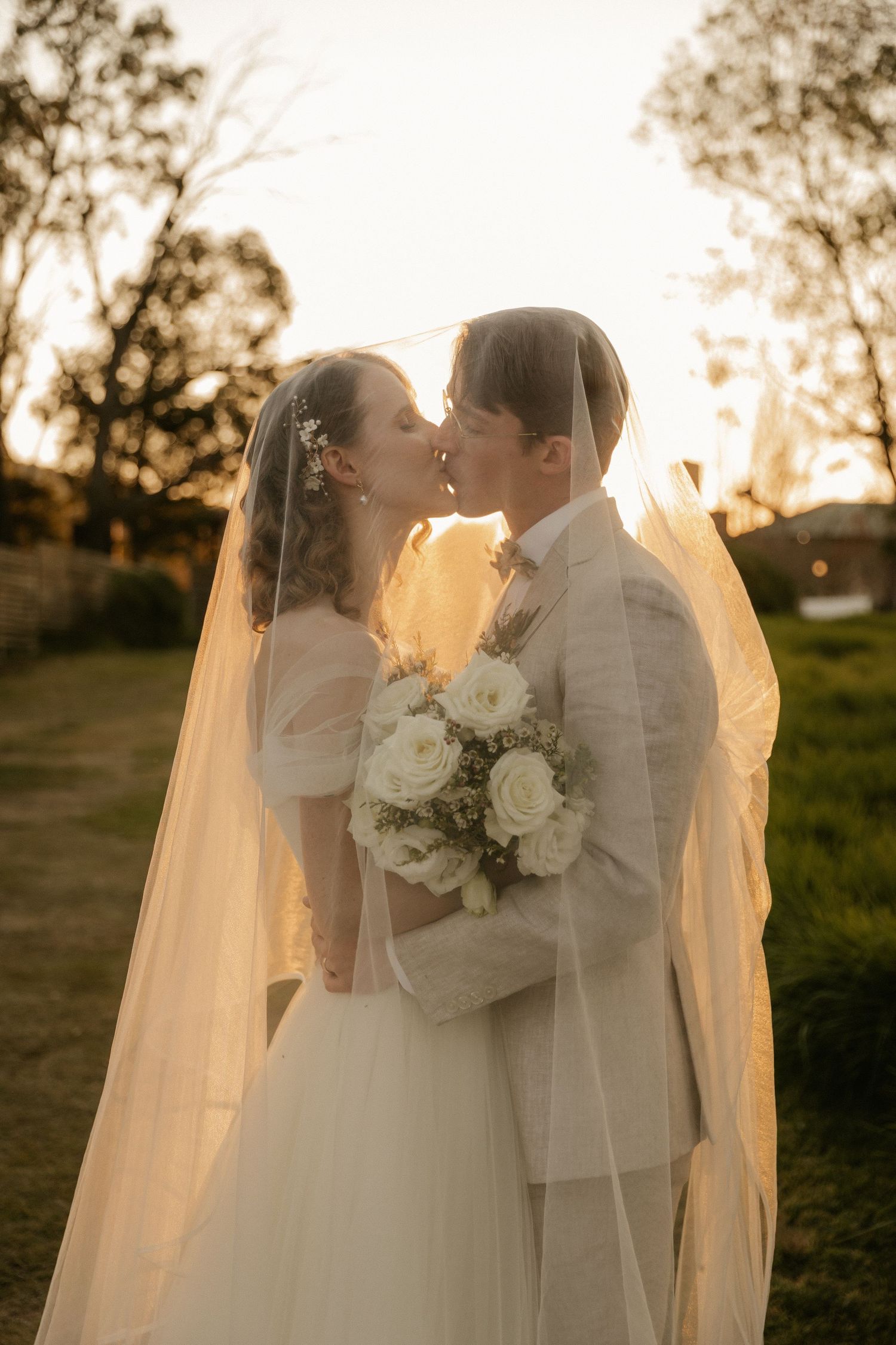 Bride and groom share a romantic sunset kiss while holding a white rose bouquet amid trees and golden light.
