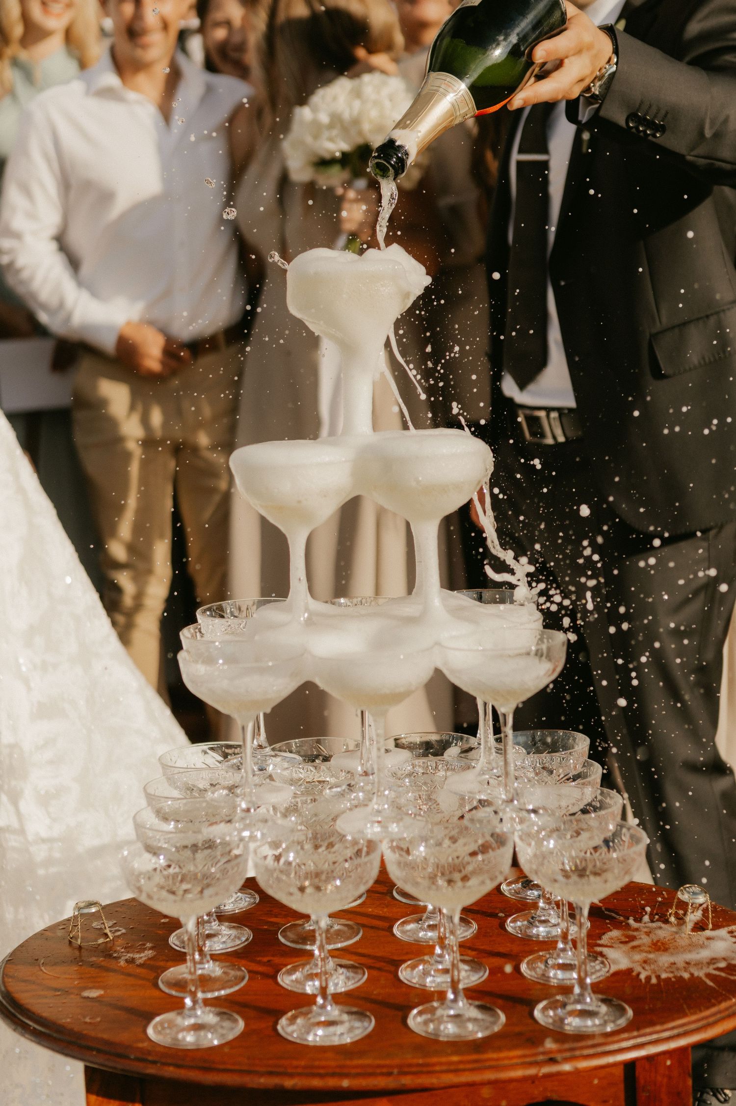 Champagne tower overflowing at a wedding reception with guests in background.