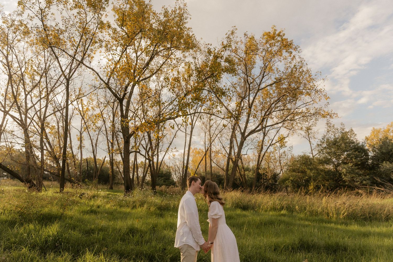 A couple stands together in a golden field at sunset with tall autumn trees behind them in soft evening light.