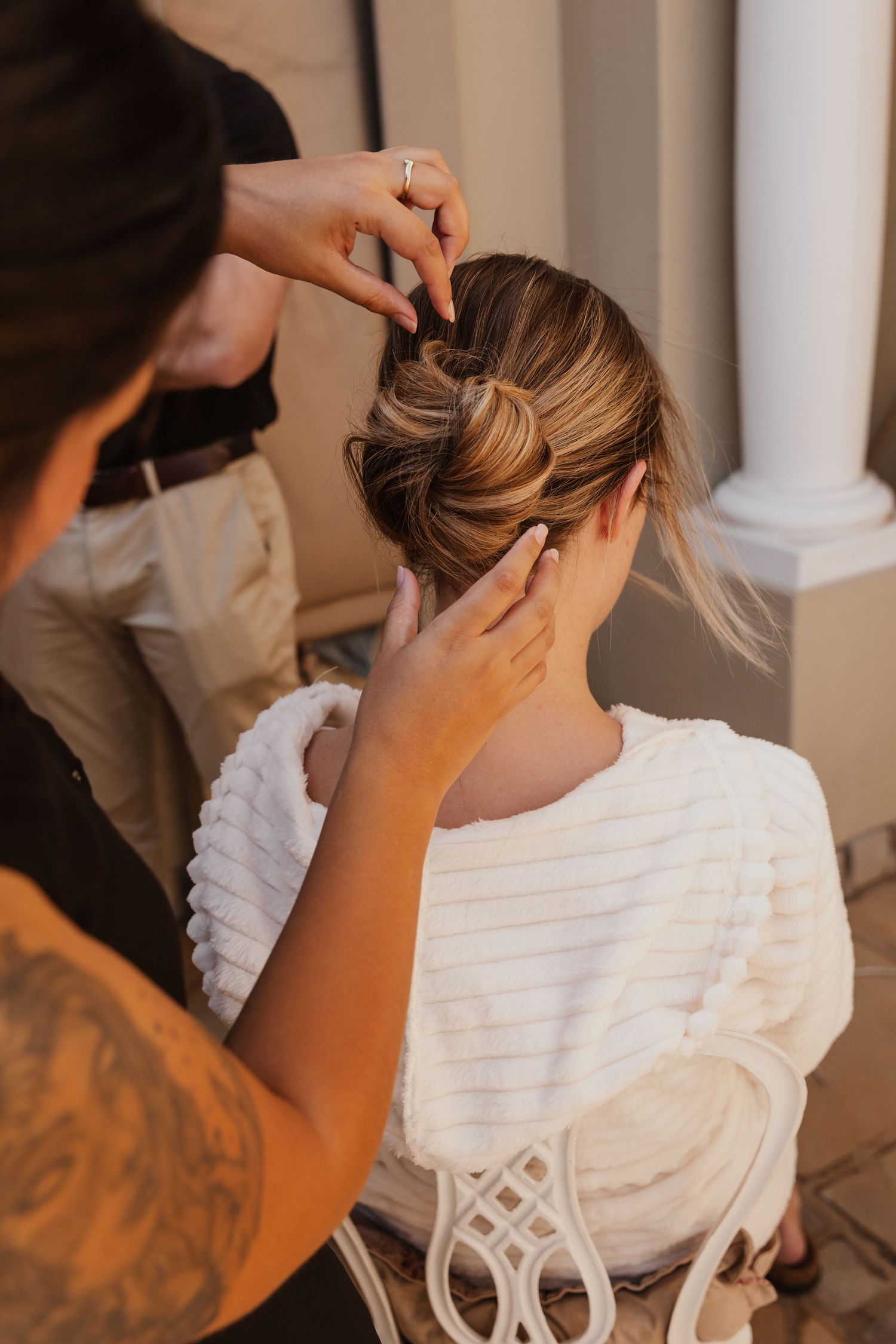 Stylist creating an intricate updo hairstyle for a bride wearing a white robe.