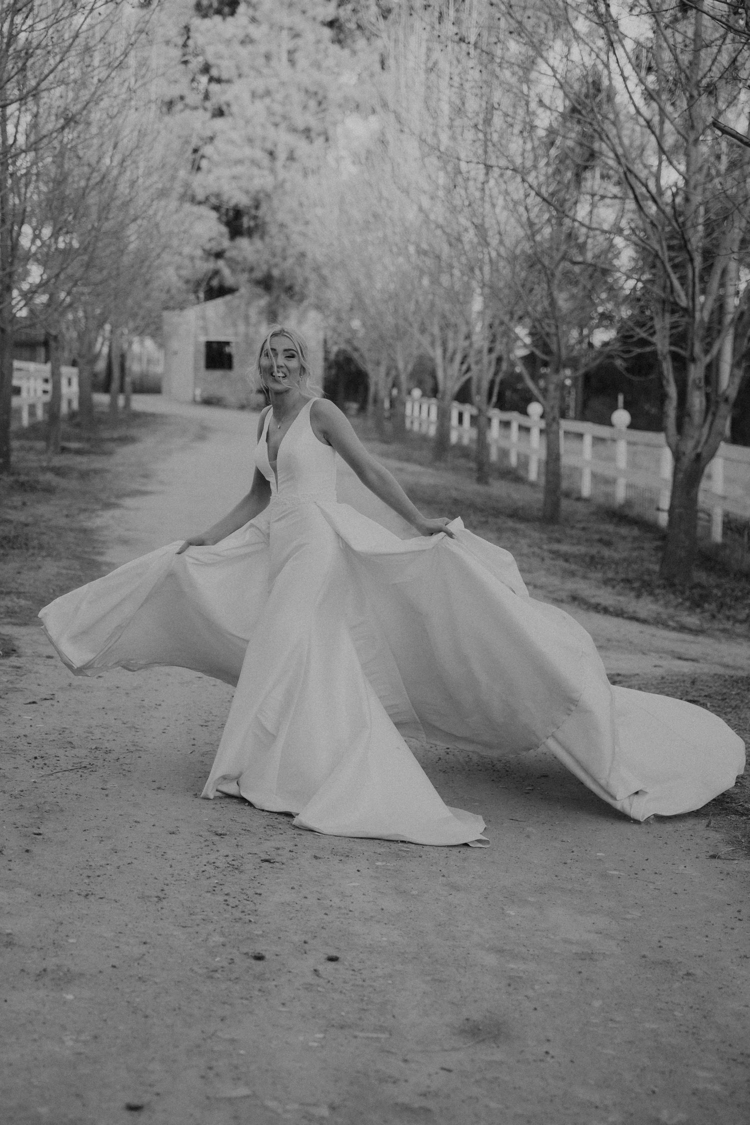 Black and white photo of a bride twirling in a flowing white wedding dress on a tree-lined path with a white fence.