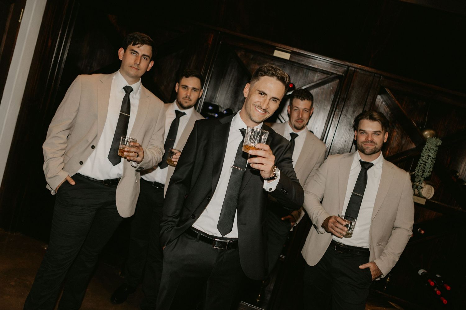 Five groomsmen in black suits and ties pose with drinks at a wedding reception venue.