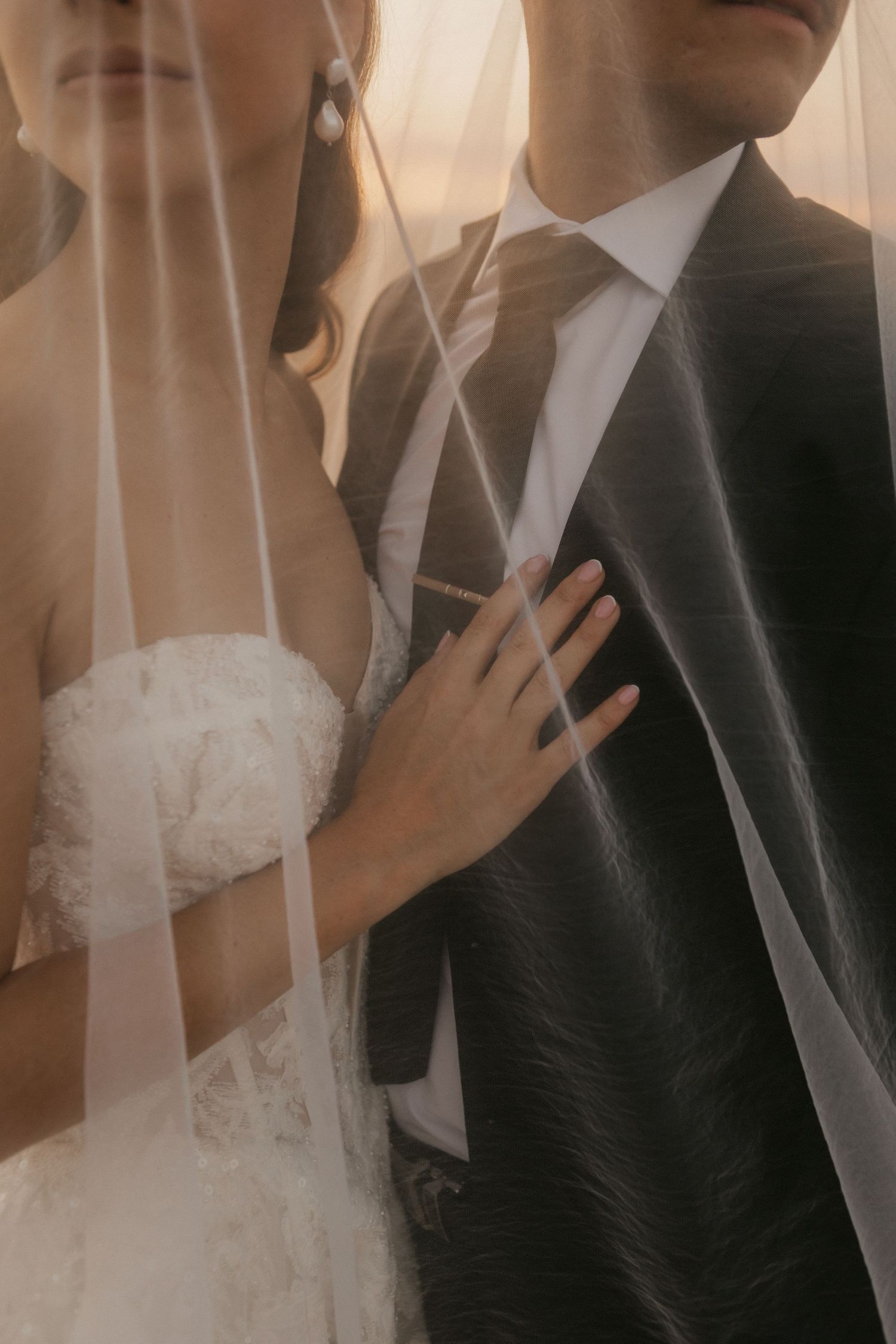 Bride in white lace gown rests hand on groom's dark suit chest, partially covered by sheer wedding veil.