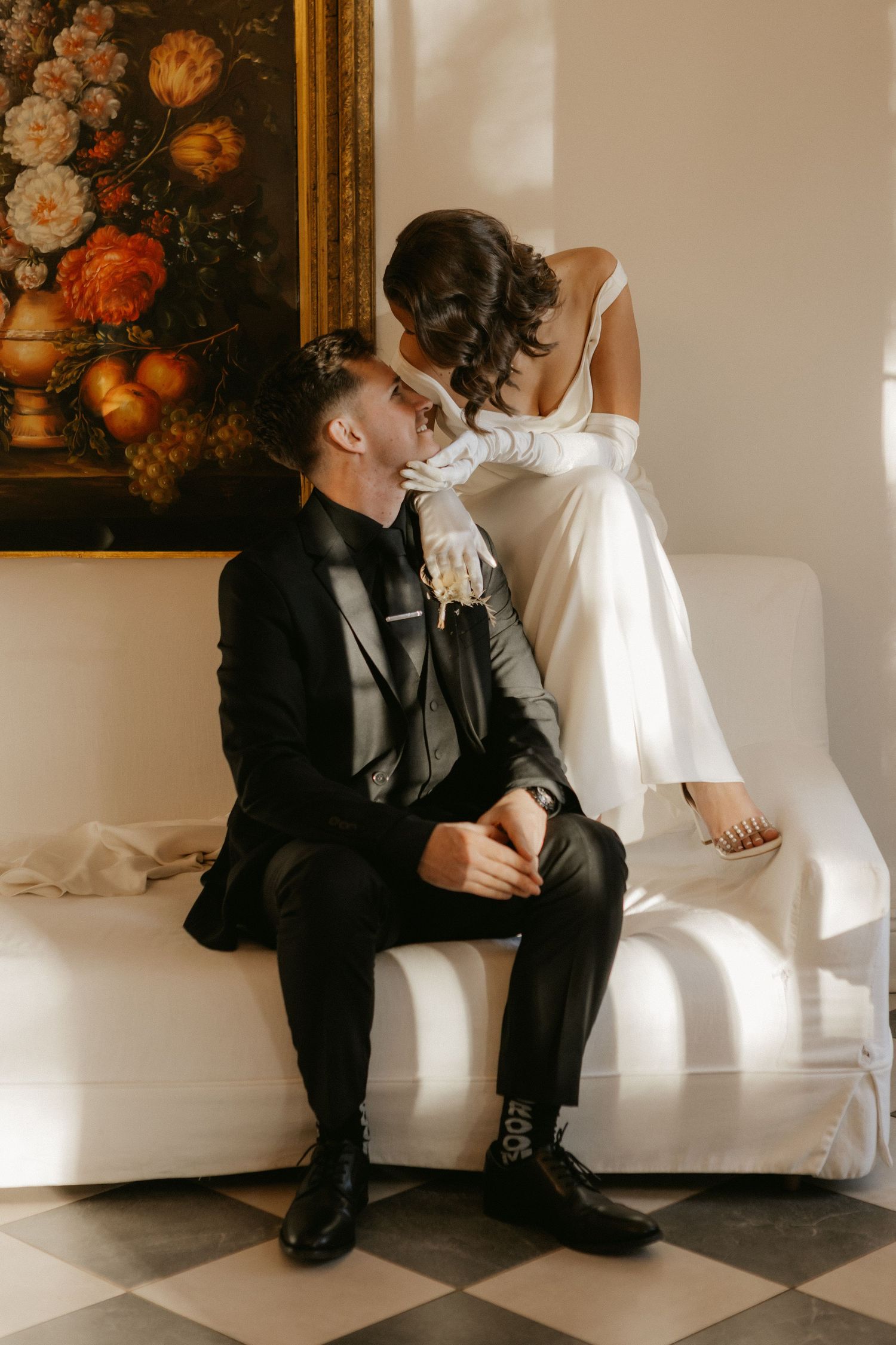 Elegant bride and groom sharing intimate moment on white sofa, with floral painting backdrop, in classic wedding attire.