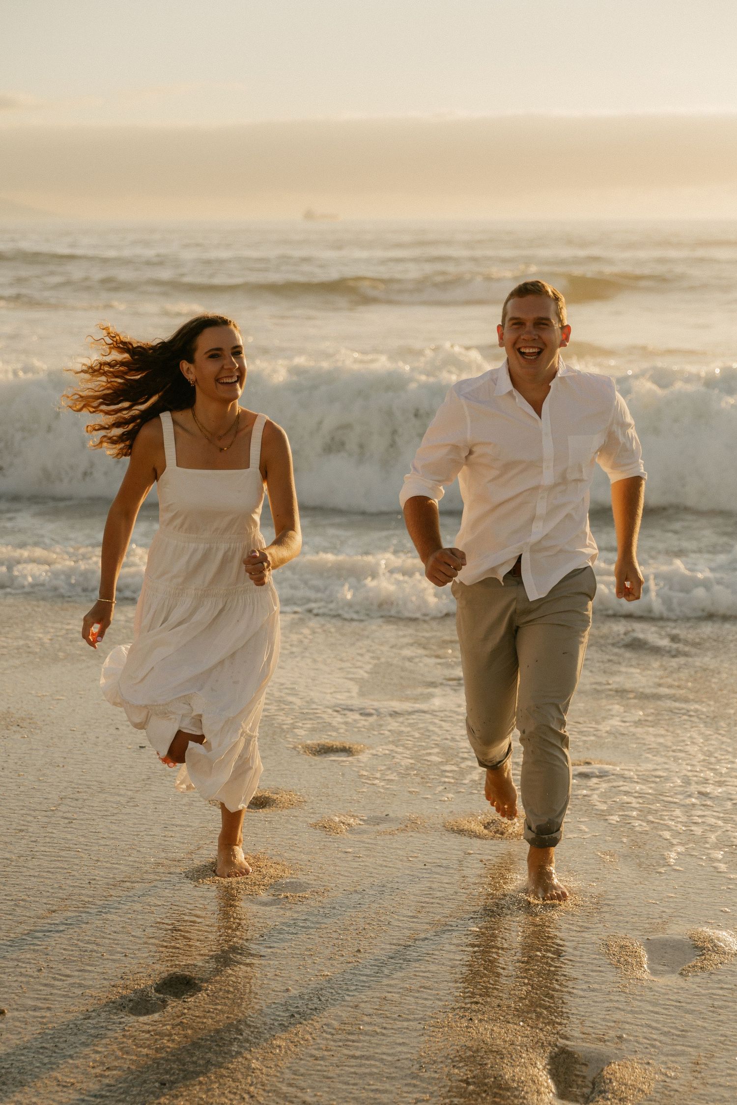 A joyful couple runs along the beach at sunset with waves crashing behind them.