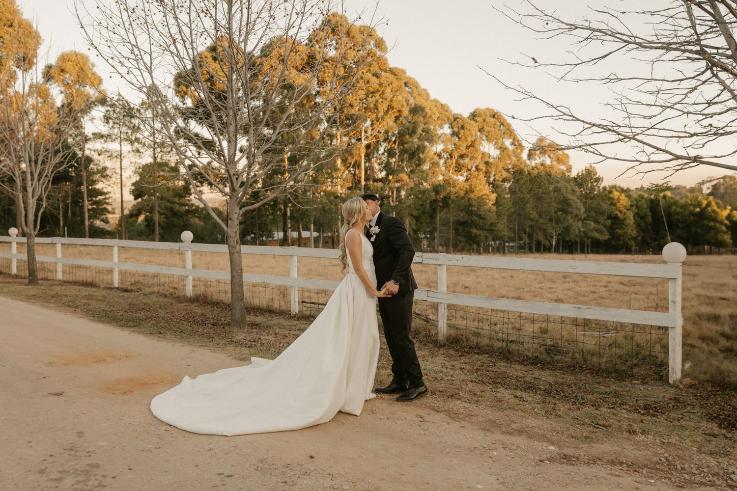A couple in wedding attire poses together on a rustic dirt path with trees and autumn foliage in the background.