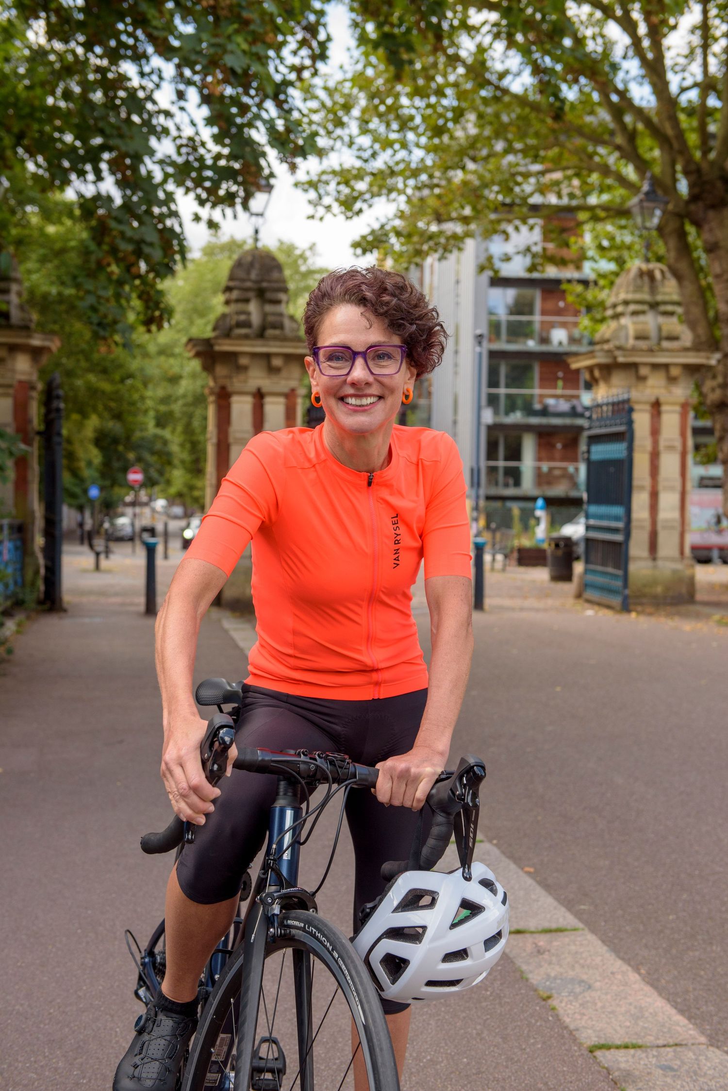 Cyclist in bright orange shirt with glasses rides a bike near decorative stone pillars on a tree-lined path.
