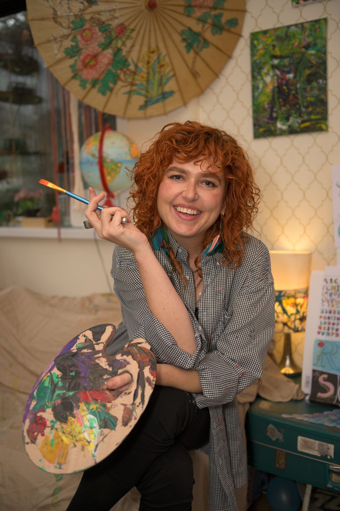 Artist in studio holding paintbrush and palette next to decorative umbrella and artwork on walls.