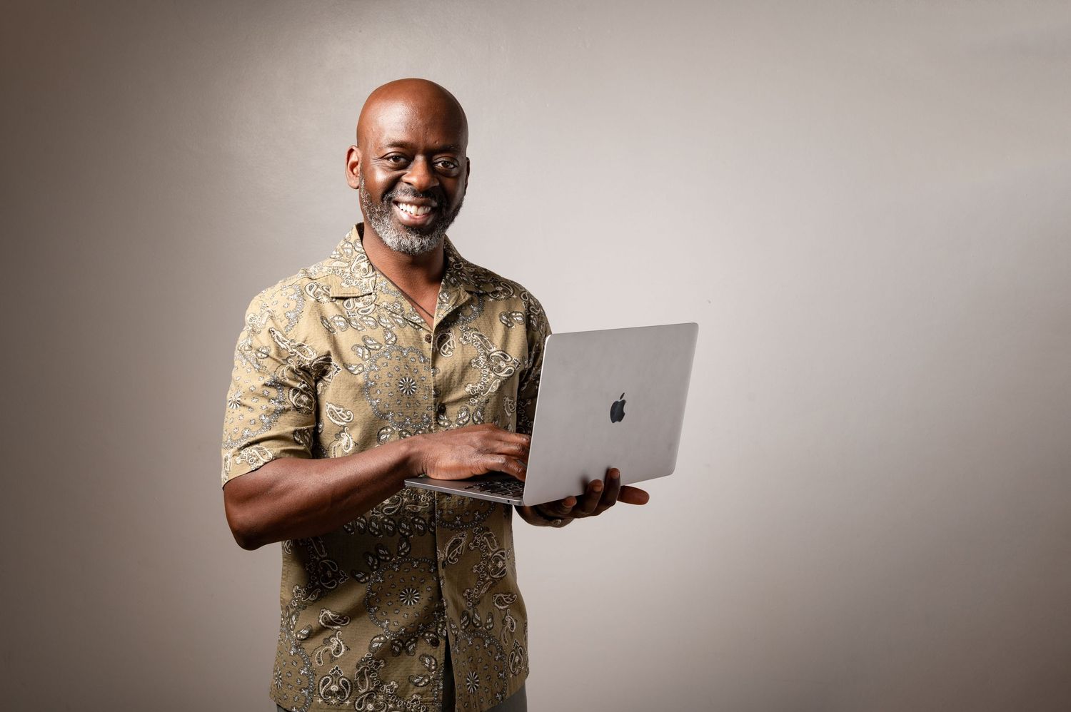 Professional looking using a MacBook laptop while smiling against a neutral background wearing a beige patterned shirt.