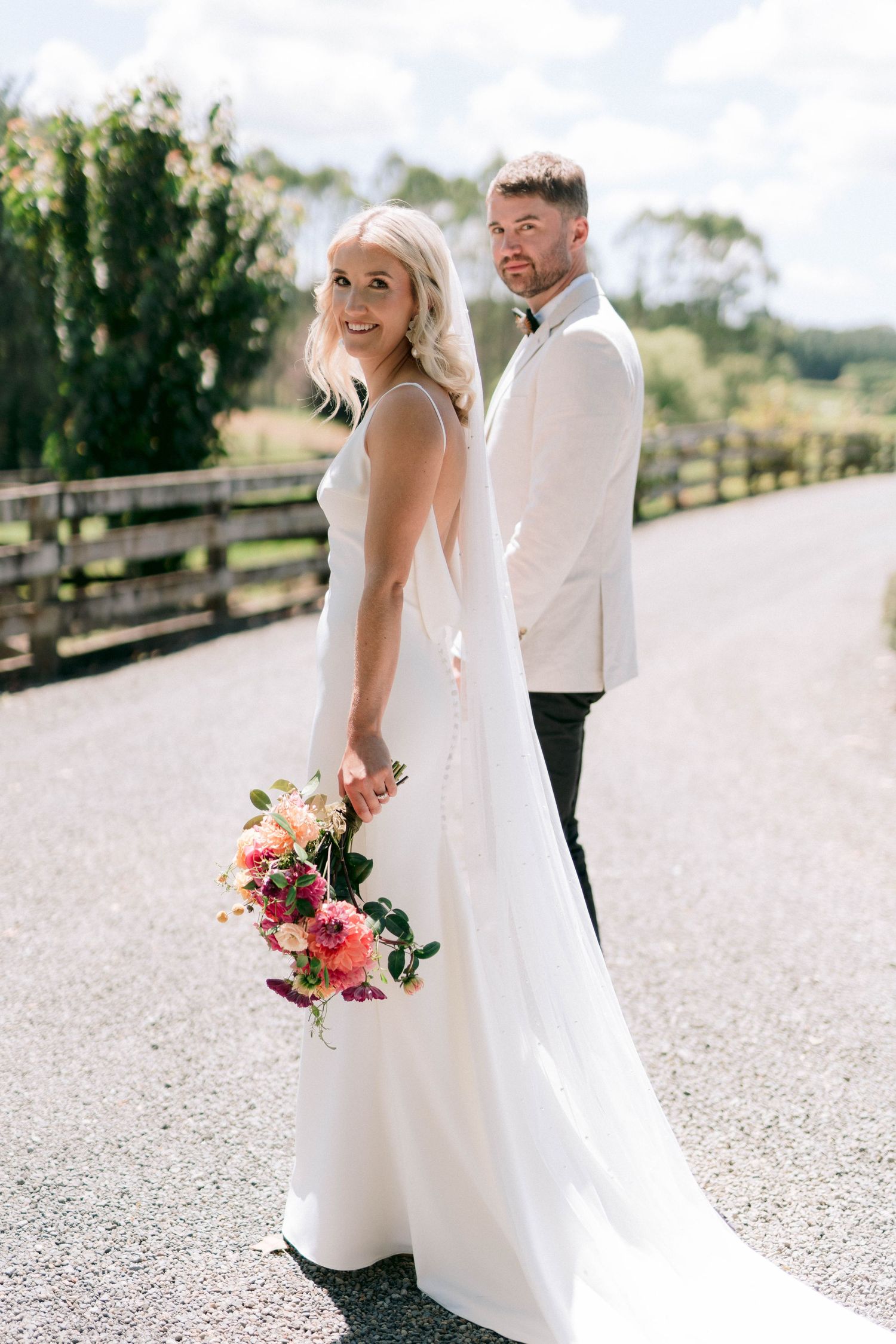 A couple in wedding attire poses together on a country road holding a colorful bouquet.
