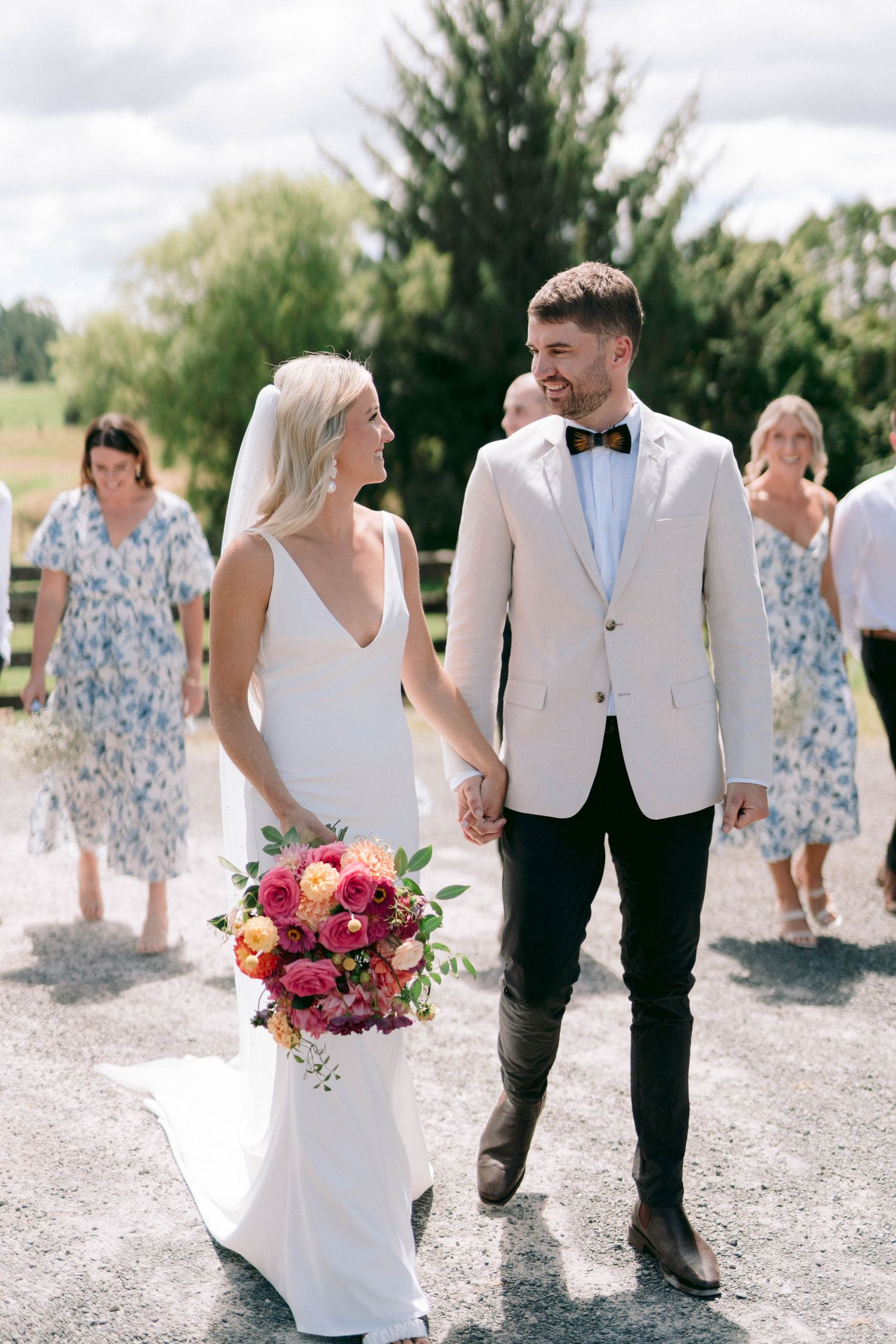 Wedding couple walks along path with guests in floral dresses following behind.